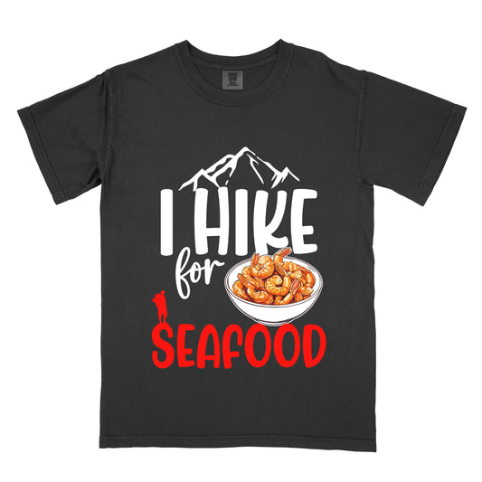 Black t-shirt with white and red text reading "I HIKE for SEAFOOD". "I HIKE" appears in large white letters with white mountain outlines above it, "for" in white script in the middle, and "SEAFOOD" in red at the bottom. Features an illustration of orange shrimp in a white bowl in the center and a small red hiker silhouette to the left of the bowl and above the word "SEAFOOD".
