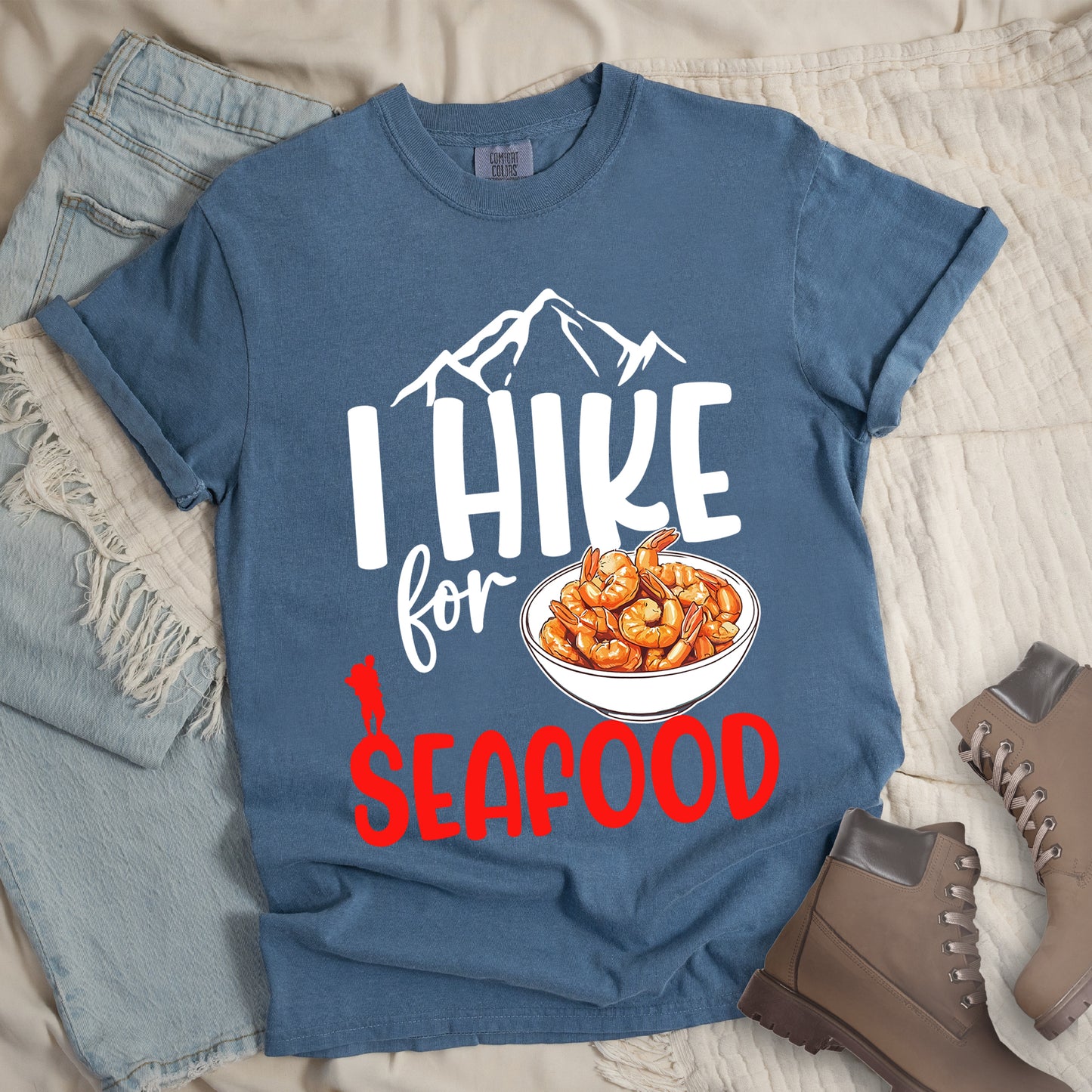 Blue Jean (Denim Blue) t-shirt with white and red text reading "I HIKE for SEAFOOD". "I HIKE" appears in large white letters with white mountain outlines above it, "for" in white script in the middle, and "SEAFOOD" in red at the bottom. Features an illustration of orange shrimp in a white bowl in the center and a small red hiker silhouette to the left of the bowl and above the word "SEAFOOD".  The shirt features a classic crew neck and casual rolled sleeves, alongside light wash denim and brown hiking boots