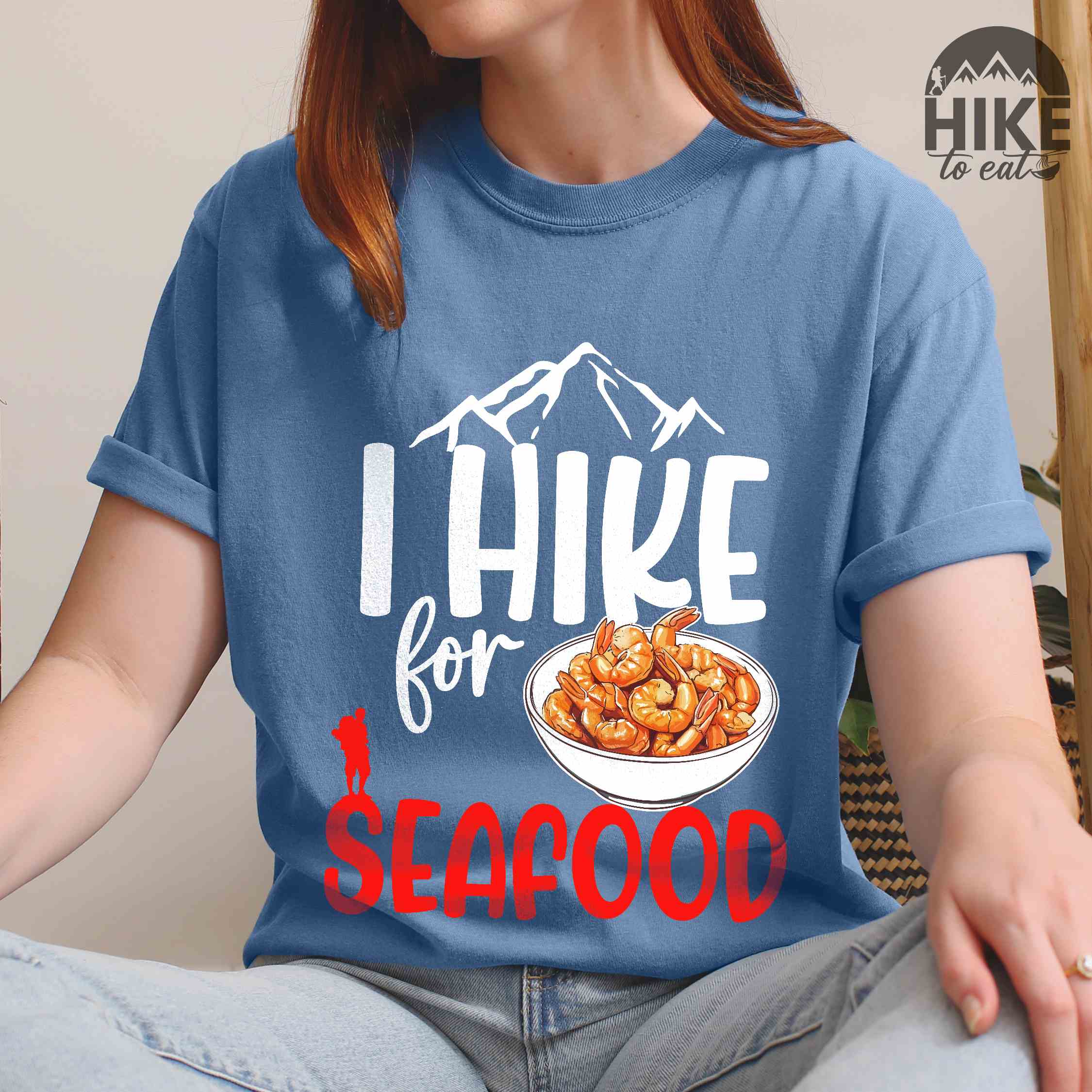 woman wearing blue jean "I hike for seafood" colored vintage tee with bold typography, illustrated shrimps on a bowl, mountains, birds, and backpackers for seafood  lovers.