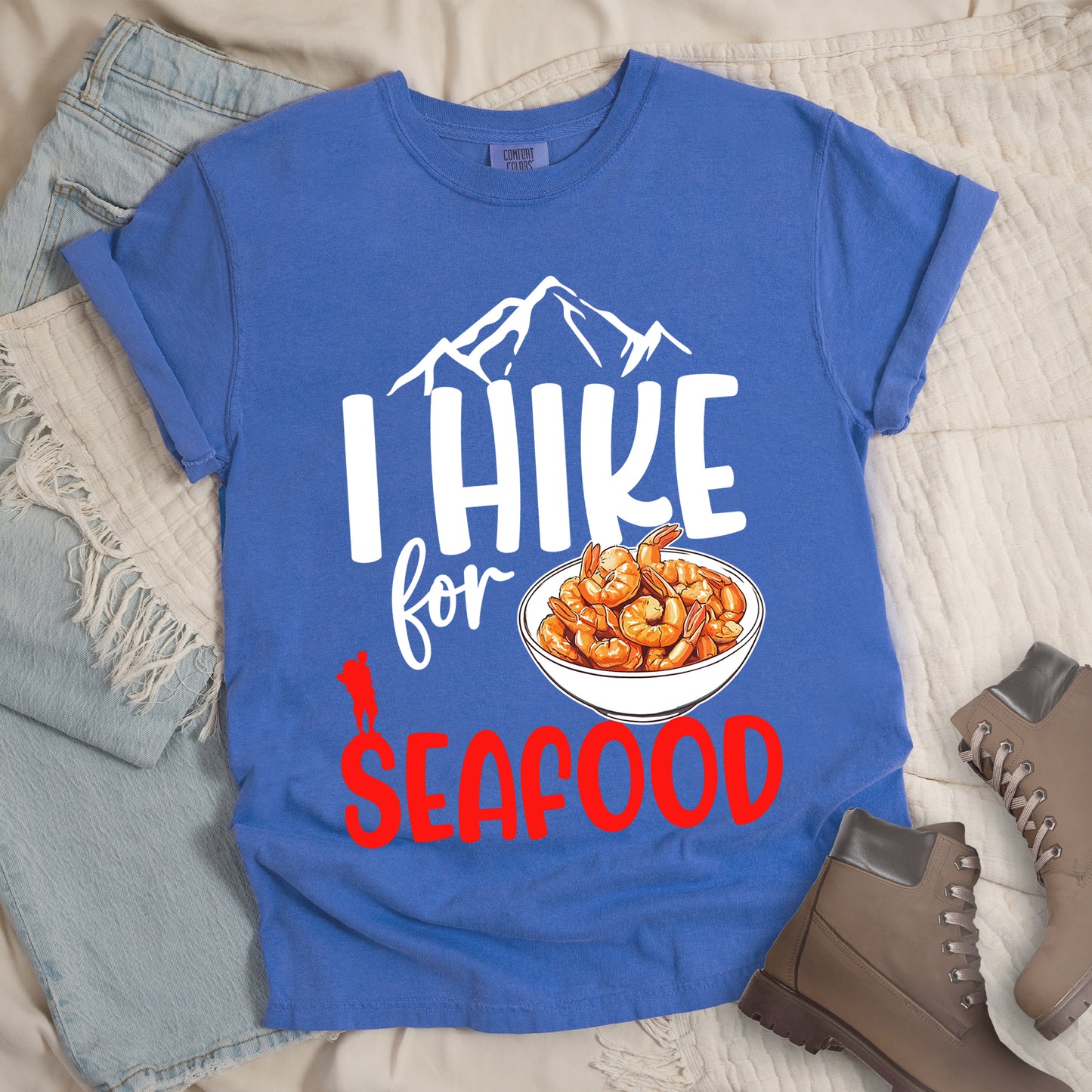 Flo Blue (Bright Blue) t-shirt with white and red text reading "I HIKE for SEAFOOD". "I HIKE" appears in large white letters with white mountain outlines above it, "for" in white script in the middle, and "SEAFOOD" in red at the bottom. Features an illustration of orange shrimp in a white bowl in the center and a small red hiker silhouette to the left of the bowl and above the word "SEAFOOD". Features a classic crew neck and casual rolled sleeves, styled alongside light wash denim and brown hiking boots. 