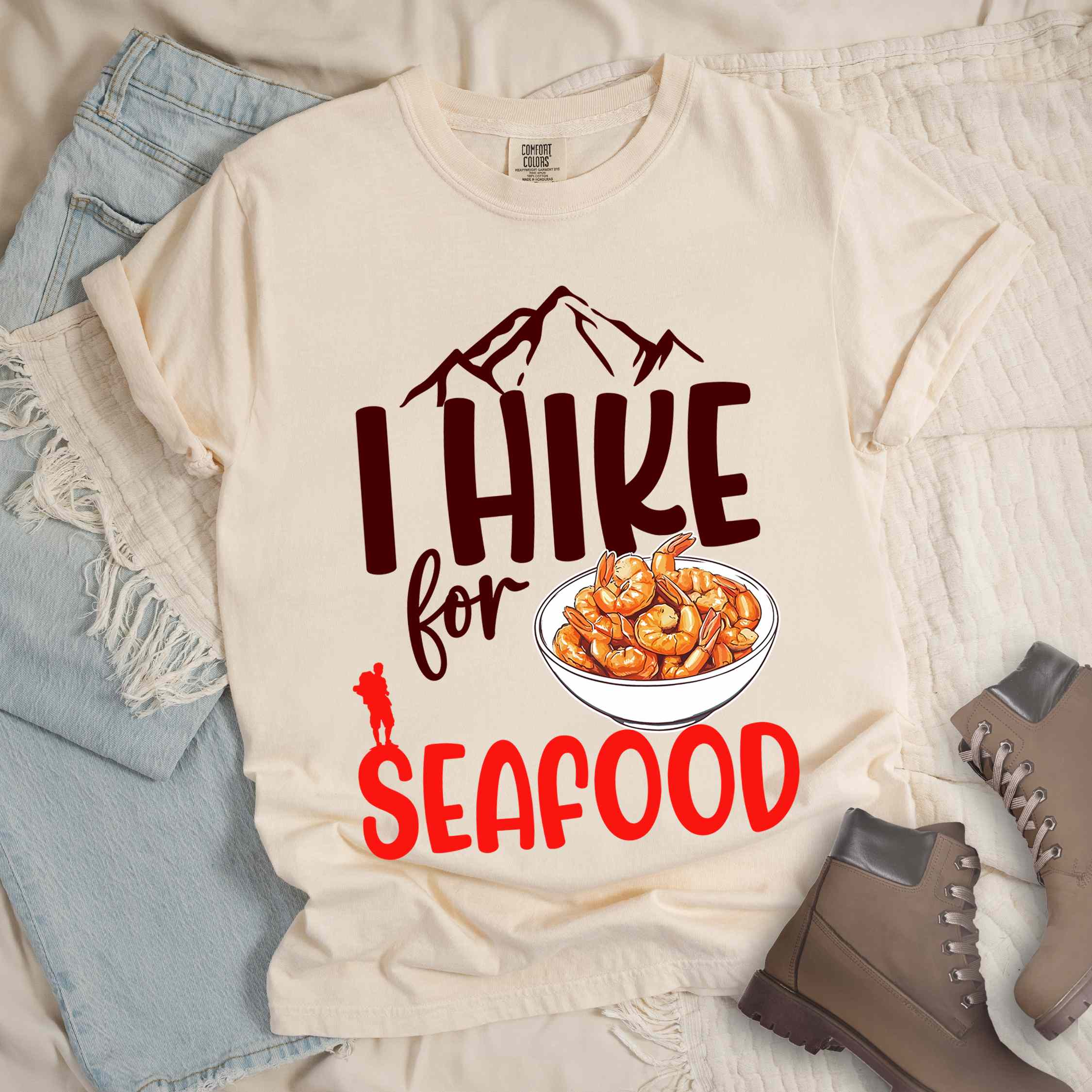 Ivory "I hike for seafood" colored vintage tee with bold typography, illustrated shrimps on a bowl, mountains, birds, and backpackers for seafood  lovers.