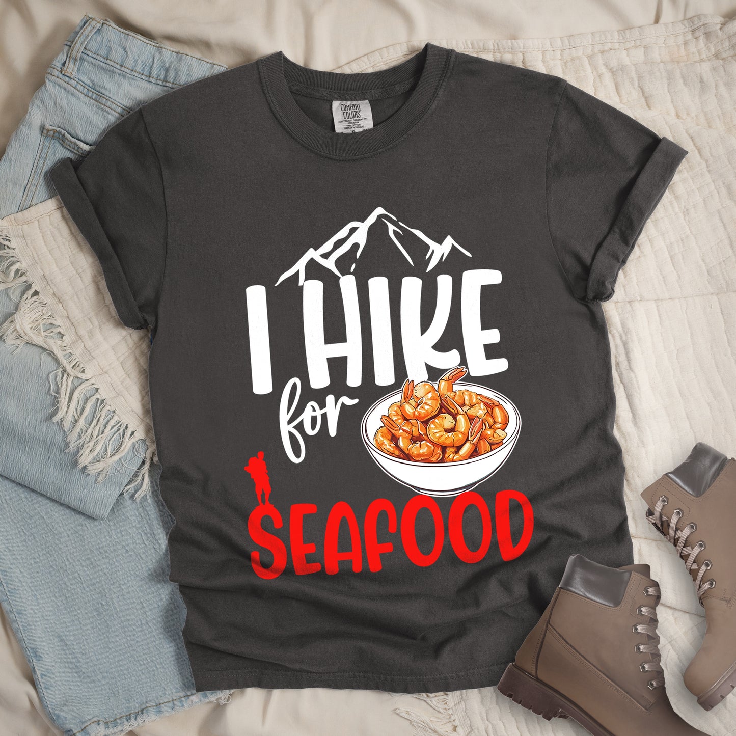 Pepper (Dark Gray) t-shirt with white and red text reading "I HIKE for SEAFOOD". "I HIKE" appears in large white letters with white mountain outlines above it, "for" in white script in the middle, and "SEAFOOD" in red at the bottom. Features an illustration of orange shrimp in a white bowl in the center and a small red hiker silhouette to the left of the bowl and above the word "SEAFOOD". Features a classic crew neck and casual rolled sleeves, styled alongside light wash denim and brown hiking boots. 