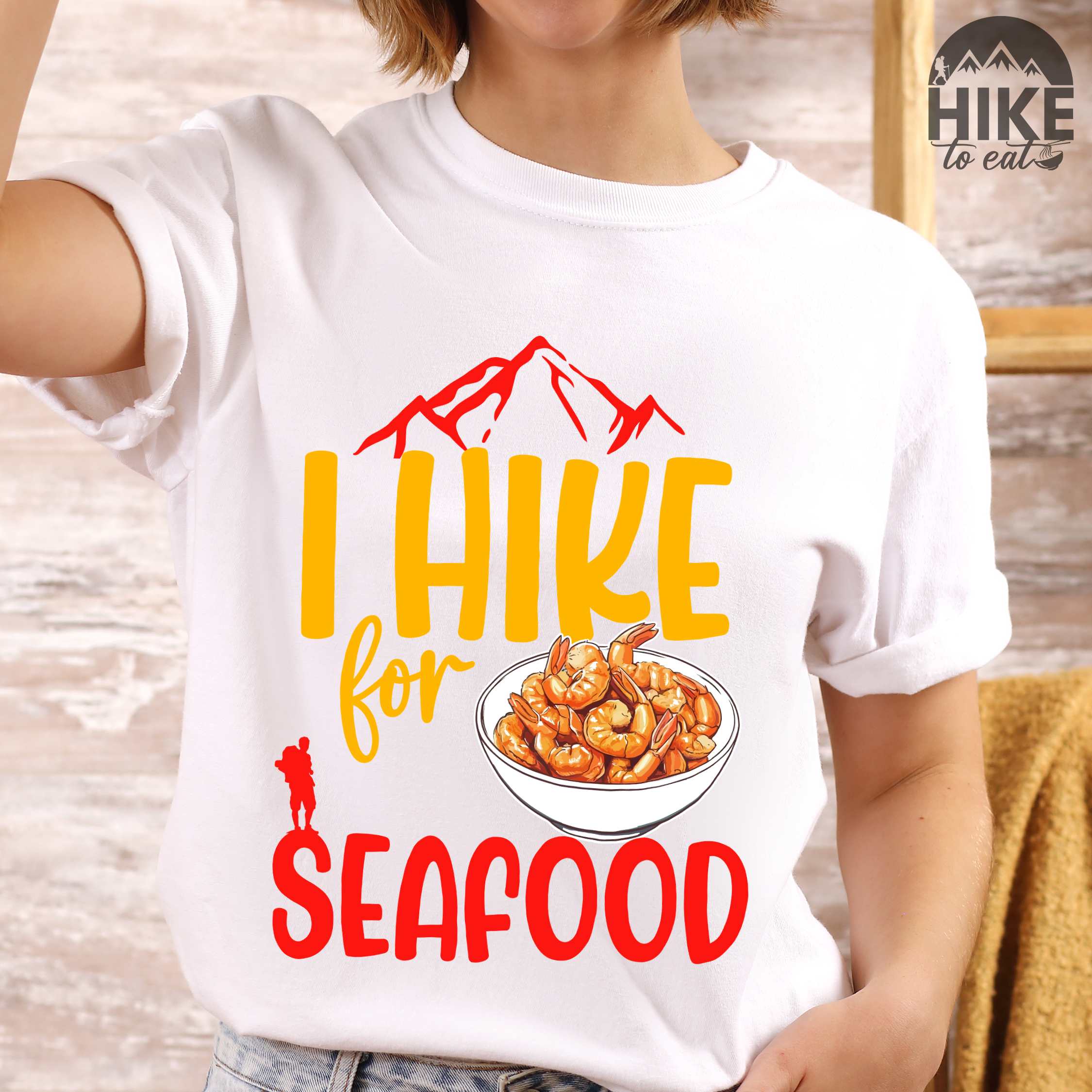 Woman wearing white "I hike for seafood" colored vintage tee with bold typography, illustrated shrimps on a bowl, mountains, birds, and backpackers for seafood  lovers.