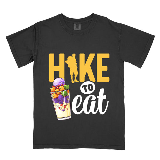 Black t-shirt with yellow and white text reading "HIKE TO eat". "HIKE" appears in large yellow letters at the top with a yellow hiker silhouette integrated into the letter "I", "TO" in black text on a white circular background in the middle, and "eat" in white script at the bottom. Features an illustration of a colorful fruit parfait/dessert (Halo-Halo) in a clear glass with purple, orange, and green fruit pieces to the left of "TO eat".