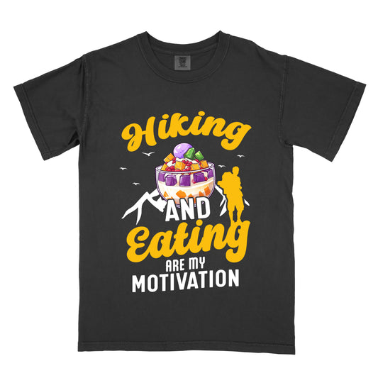 Black t-shirt with yellow and white text reading "Hiking AND Eating ARE MY MOTIVATION". "Hiking" appears in yellow script at the top, "AND" in white in the middle, "Eating" in yellow script below it, and "ARE MY MOTIVATION" in white at the bottom. Features an illustration of a colorful dessert with purple, orange and red fruit pieces (Halo-Halo) in the center, with ube ice cream on top, white mountain outlines, small white birds, and a yellow silhouette of a hiker to the right of "AND".