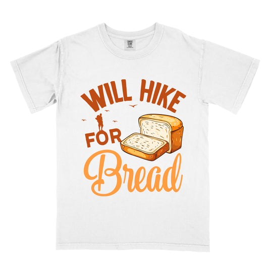 White t-shirt with brown and orange text reading "WILL HIKE FOR Bread". "WILL HIKE" appears in brown arched text at the top, "FOR" in brown below it, and "Bread" in orange script at the bottom. Features an illustration of a loaf of bread with one slice cut off to the right of "FOR", small brown birds flying, and a brown silhouette of a hiker above "FOR". The design humorously suggests hiking to earn bread as a reward.