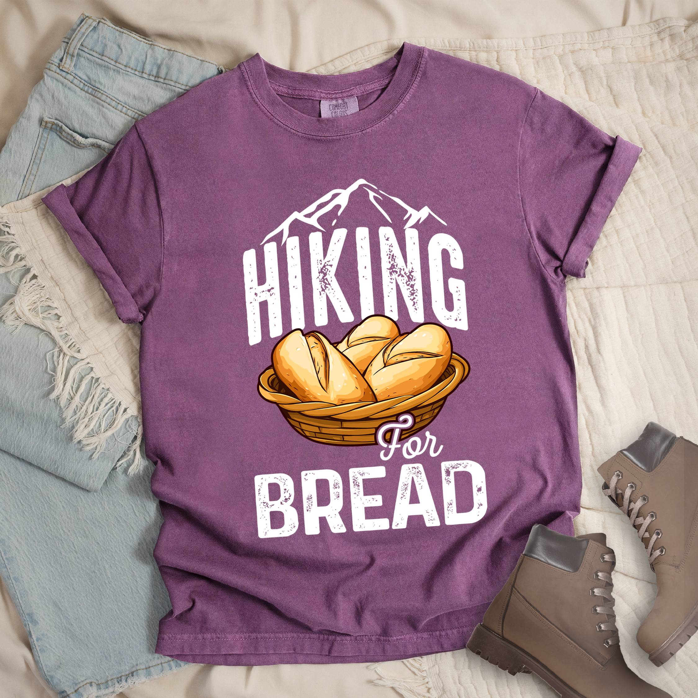 Dark pink t-shirt with white distressed text reading "HIKING For BREAD". "HIKING" appears in large white distressed letters at the top with white mountain outlines above it, "For" in small gray script in the middle, and "BREAD" in large white distressed letters at the bottom. Features an illustration of bread rolls in a woven basket between the words "HIKING" and "BREAD". Features a classic crew neck and casual rolled sleeves, styled alongside light wash denim and brown hiking boots. 
