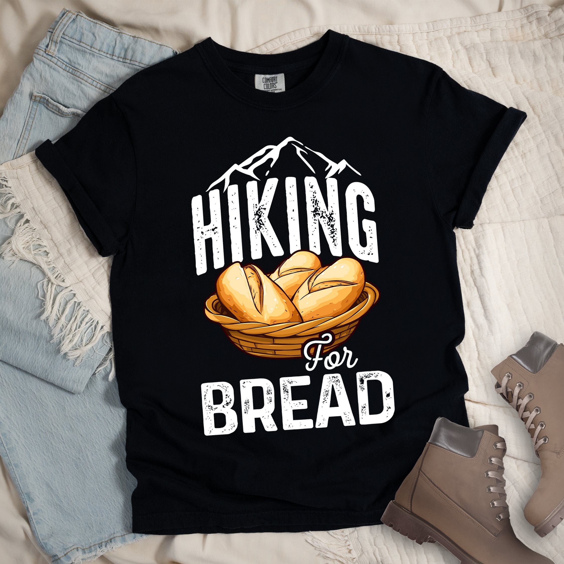Black t-shirt with white distressed text reading "HIKING For BREAD". "HIKING" appears in large white distressed letters at the top with white mountain outlines above it, "For" in small gray script in the middle, and "BREAD" in large white distressed letters at the bottom. Features an illustration of bread rolls in a woven basket between the words "HIKING" and "BREAD". Features a classic crew neck and casual rolled sleeves, styled alongside light wash denim and brown hiking boots. 
