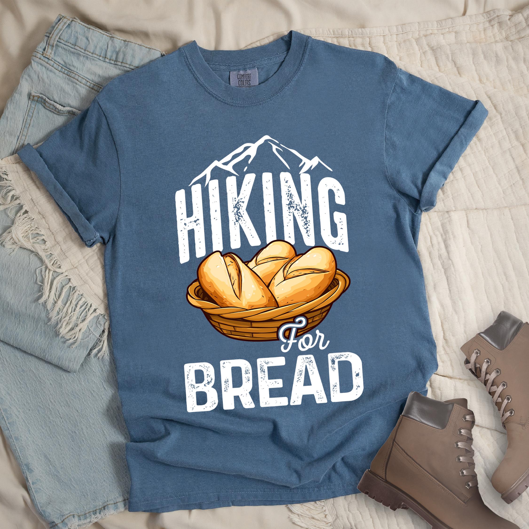 Dark blue t-shirt with white distressed text reading "HIKING For BREAD". "HIKING" appears in large white distressed letters at the top with white mountain outlines above it, "For" in small gray script in the middle, and "BREAD" in large white distressed letters at the bottom. Features an illustration of bread rolls in a woven basket between the words "HIKING" and "BREAD". Features a classic crew neck and casual rolled sleeves, styled alongside light wash denim and brown hiking boots. 