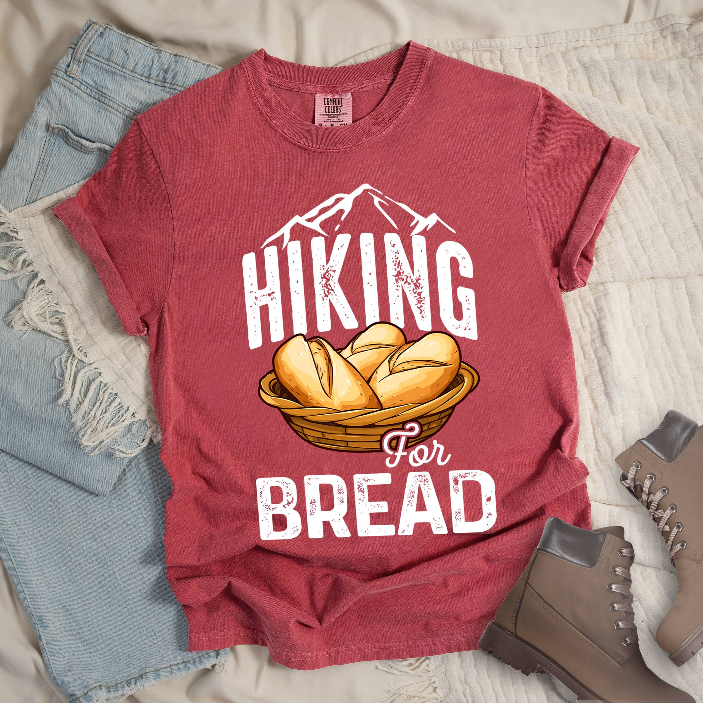 Red t-shirt with white distressed text reading "HIKING For BREAD". "HIKING" appears in large white distressed letters at the top with white mountain outlines above it, "For" in small gray script in the middle, and "BREAD" in large white distressed letters at the bottom. Features an illustration of bread rolls in a woven basket between the words "HIKING" and "BREAD". Features a classic crew neck and casual rolled sleeves, styled alongside light wash denim and brown hiking boots. 