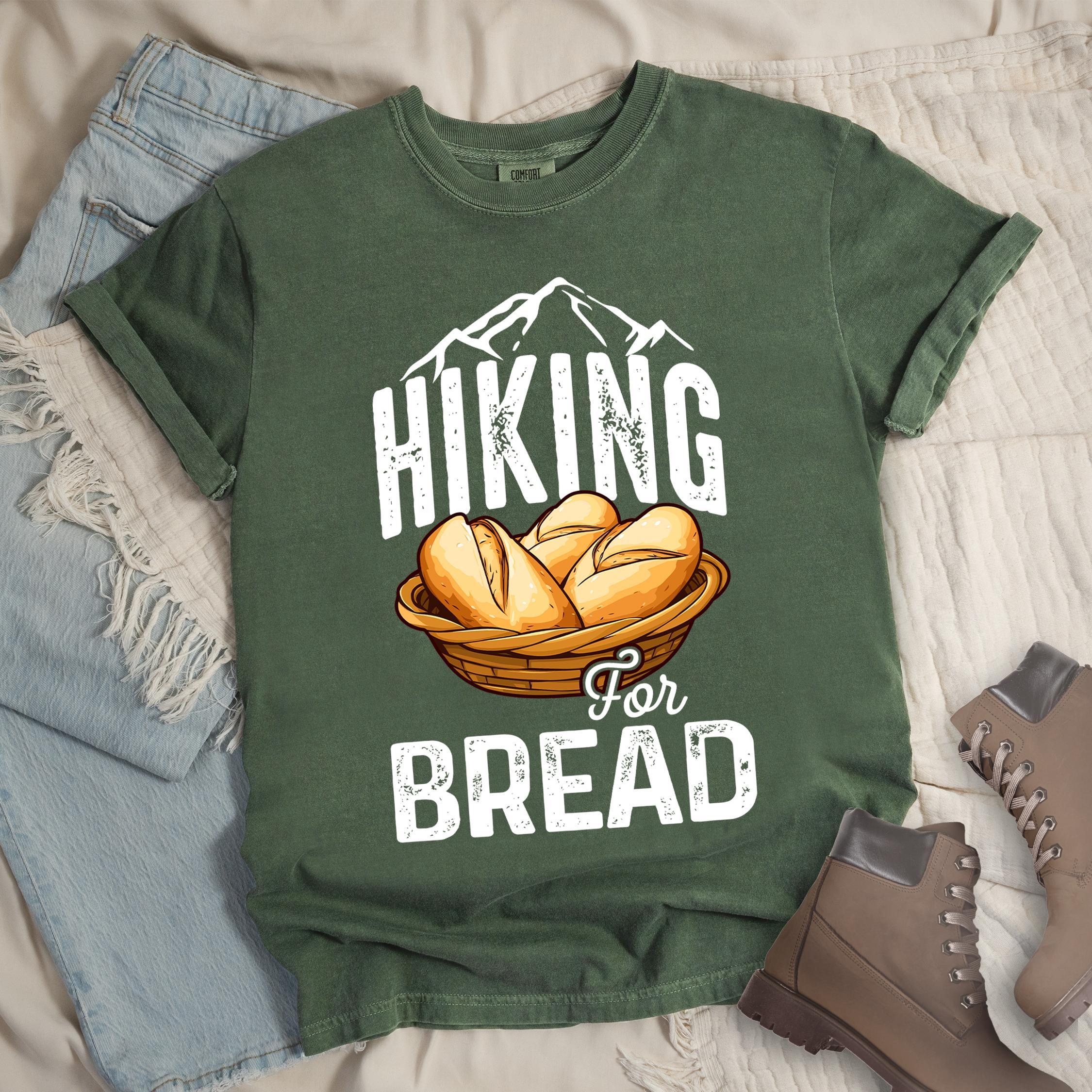 Dark green t-shirt with white distressed text reading "HIKING For BREAD". "HIKING" appears in large white distressed letters at the top with white mountain outlines above it, "For" in small gray script in the middle, and "BREAD" in large white distressed letters at the bottom. Features an illustration of bread rolls in a woven basket between the words "HIKING" and "BREAD". Features a classic crew neck and casual rolled sleeves, styled alongside light wash denim and brown hiking boots. 