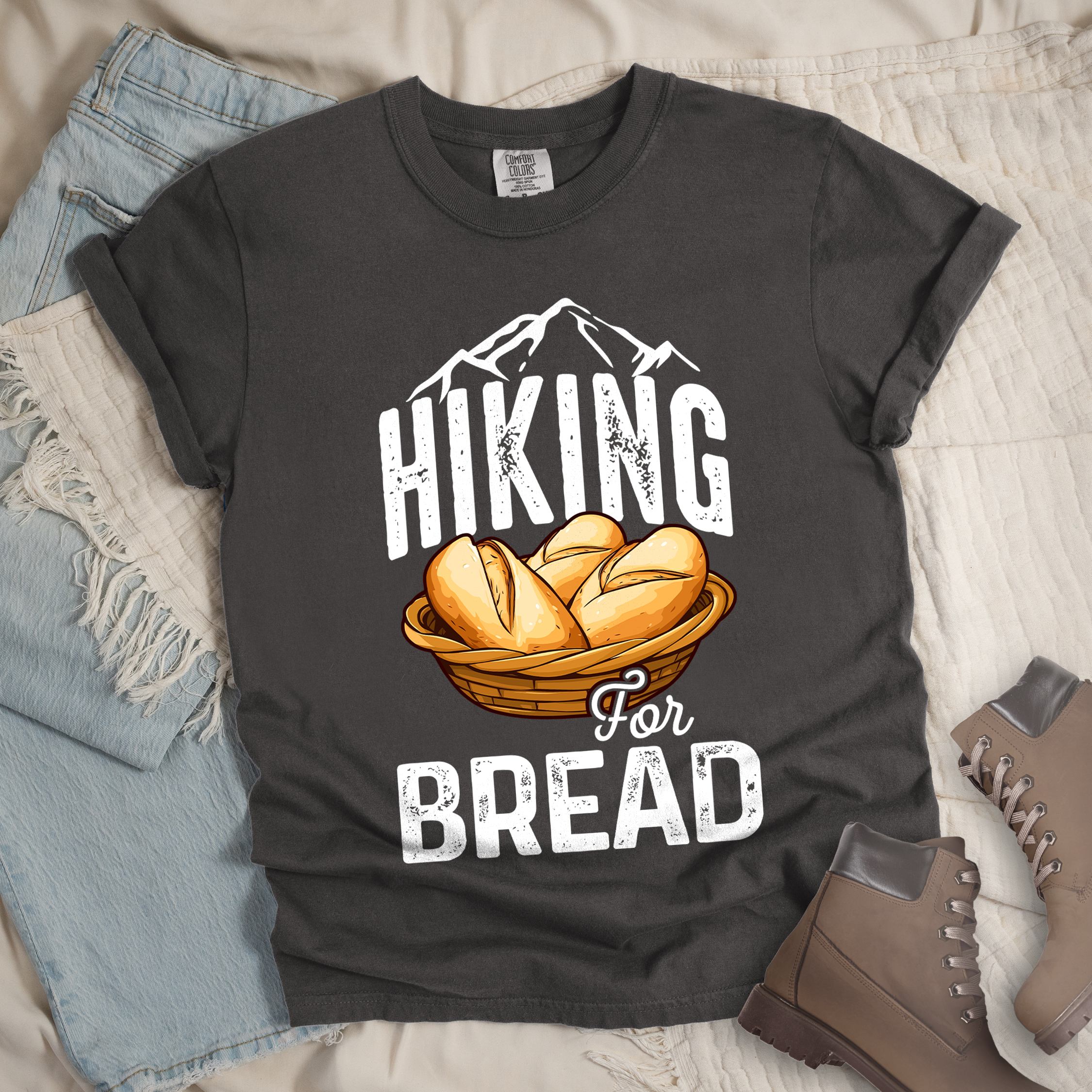 Dark grey t-shirt with white distressed text reading "HIKING For BREAD". "HIKING" appears in large white distressed letters at the top with white mountain outlines above it, "For" in small gray script in the middle, and "BREAD" in large white distressed letters at the bottom. Features an illustration of bread rolls in a woven basket between the words "HIKING" and "BREAD". Features a classic crew neck and casual rolled sleeves, styled alongside light wash denim and brown hiking boots. 