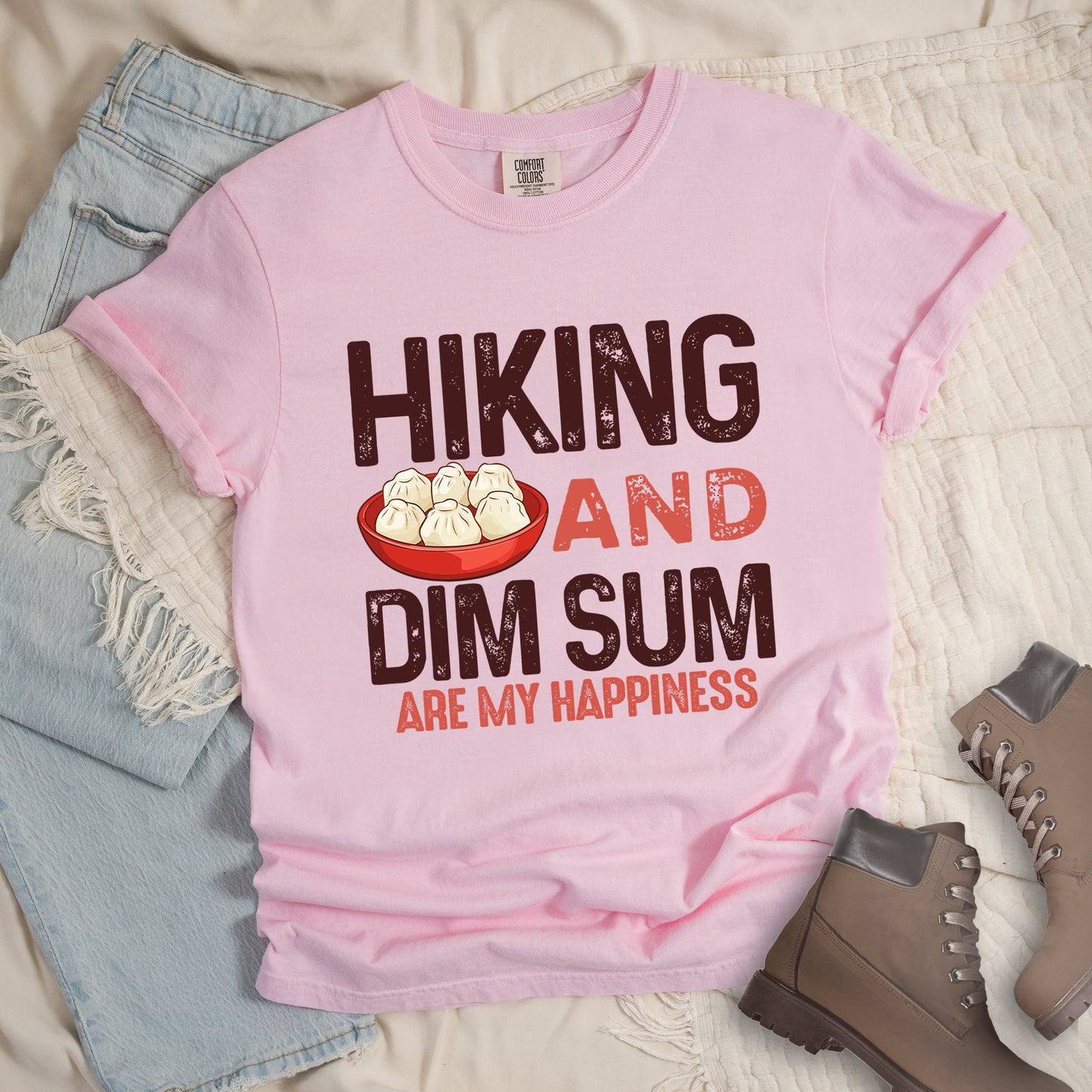 Blossom (Light Pink) t-shirt with text reading "HIKING AND DIM SUM ARE MY HAPPINESS". "HIKING" appears in large brown distressed letters at the top, "AND" in red distressed text in the middle, "DIM SUM" in large brown distressed letters below it, and "ARE MY HAPPINESS" in smaller red text at the bottom. Features an illustration of six steamed dumplings in a red bowl between "HIKING" and "AND". Features a classic crew neck and casual rolled sleeves, styled alongside light wash denim and brown hiking boots. 