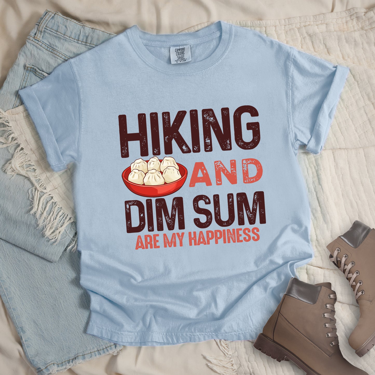Chambray (Light Blue) t-shirt with text reading "HIKING AND DIM SUM ARE MY HAPPINESS". "HIKING" appears in large brown distressed letters at the top, "AND" in red distressed text in the middle, "DIM SUM" in large brown distressed letters below it, and "ARE MY HAPPINESS" in smaller red text at the bottom. Features an illustration of six steamed dumplings in a red bowl between "HIKING" and "AND". Features a classic crew neck and casual rolled sleeves, styled alongside light wash denim and brown hiking boots. 