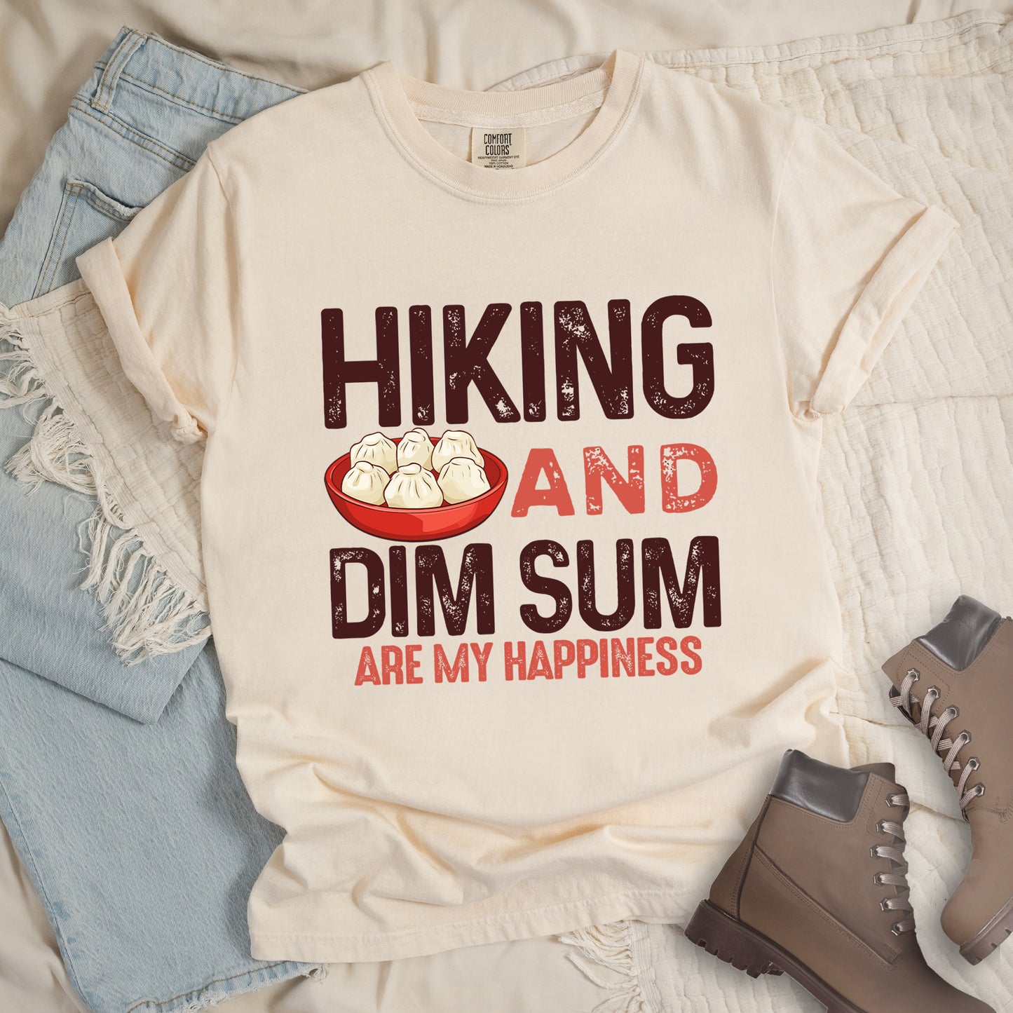 Ivory (Cream White) t-shirt with text  "HIKING AND DIM SUM ARE MY HAPPINESS". "HIKING" appears in large brown distressed letters at the top, "AND" in red distressed text in the middle, "DIM SUM" in large brown distressed letters below it, and "ARE MY HAPPINESS" in smaller red text at the bottom. Features an illustration of six steamed dumplings in a red bowl between "HIKING" and "AND". Features a classic crew neck and casual rolled sleeves, styled alongside light wash denim and brown hiking boots. 