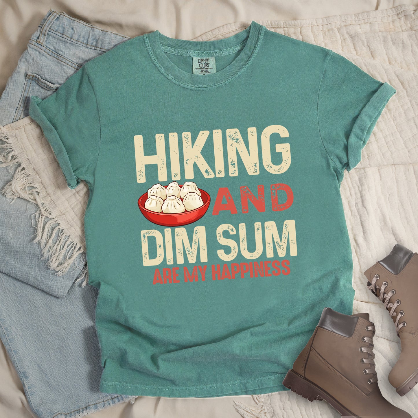 Light Green t-shirt with text "HIKING AND DIM SUM ARE MY HAPPINESS". "HIKING" appears in large beige distressed letters at the top, "AND" in red distressed text in the middle, "DIM SUM" in large beige distressed letters below it, and "ARE MY HAPPINESS" in smaller red text at the bottom. Features an illustration of six steamed dumplings in a red bowl between "HIKING" and "AND". Features a classic crew neck and casual rolled sleeves, styled alongside light wash denim and brown hiking boots. 