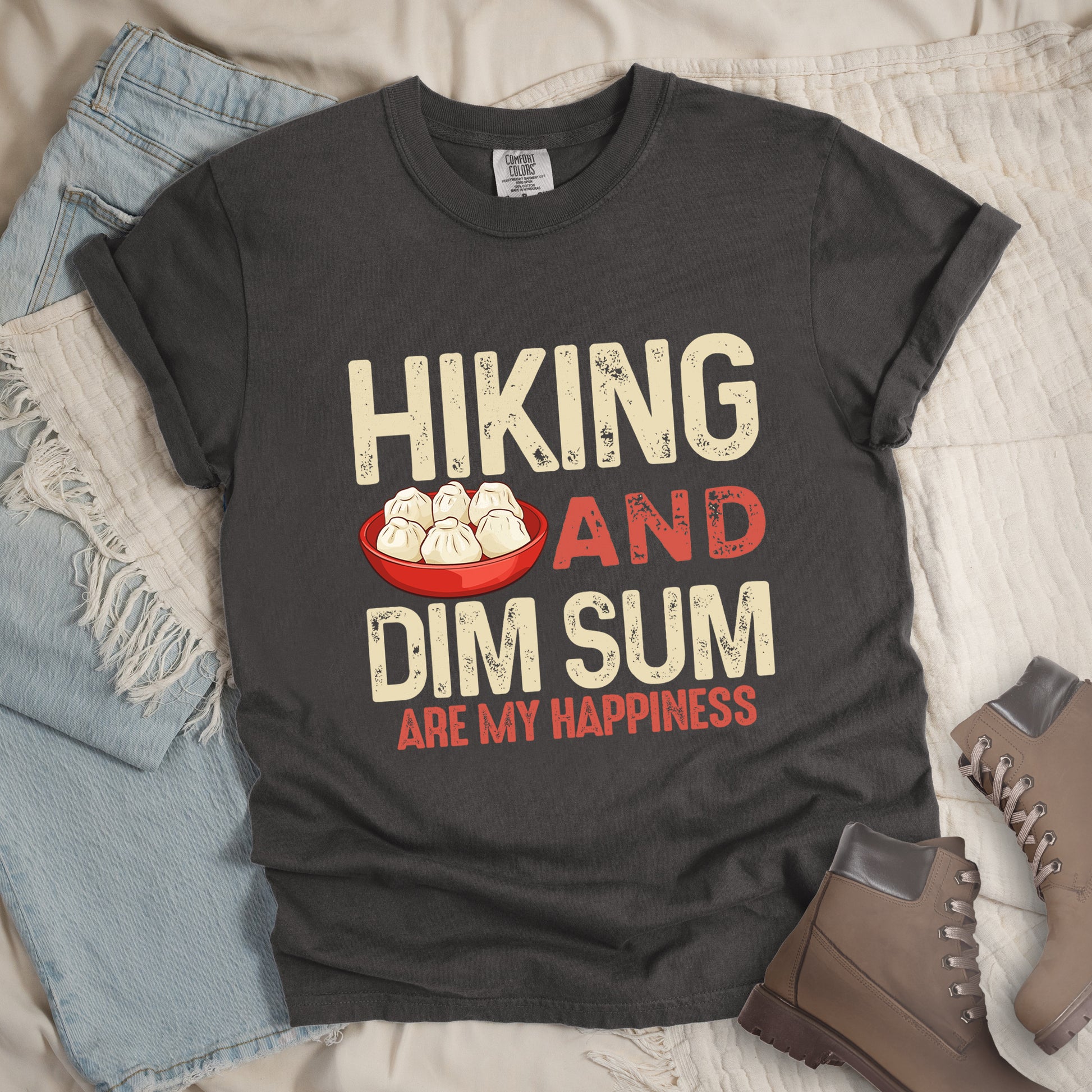 Pepper (Dark Gray) t-shirt with text "HIKING AND DIM SUM ARE MY HAPPINESS". "HIKING" appears in large beige distressed letters at the top, "AND" in red distressed text in the middle, "DIM SUM" in large beige distressed letters below it, and "ARE MY HAPPINESS" in smaller red text at the bottom. Features an illustration of six steamed dumplings in a red bowl between "HIKING" and "AND". Features a classic crew neck and casual rolled sleeves, styled alongside light wash denim and brown hiking boots. 