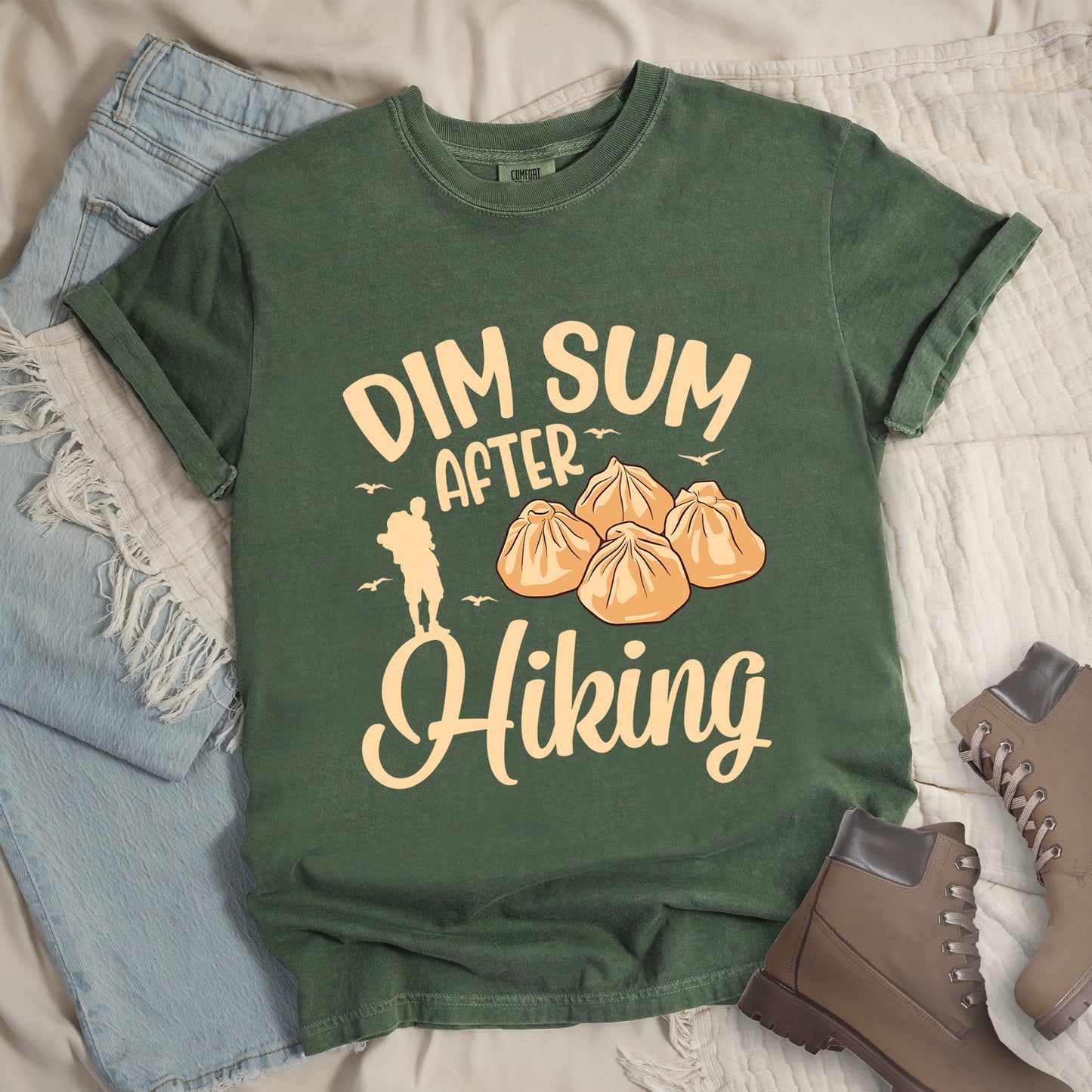 Moss (Dark Green) t-shirt with beige text reading "DIM SUM AFTER Hiking". "DIM SUM" appears in large beige letters at the top, "AFTER" in smaller beige text in the middle, and "Hiking" in beige script at the bottom. Features an illustration of four steamed dumplings in the center, small beige flying birds, and a beige silhouette of a hiker to the left of "AFTER". Features a classic crew neck and casual rolled sleeves, styled alongside light wash denim and brown hiking boots. 