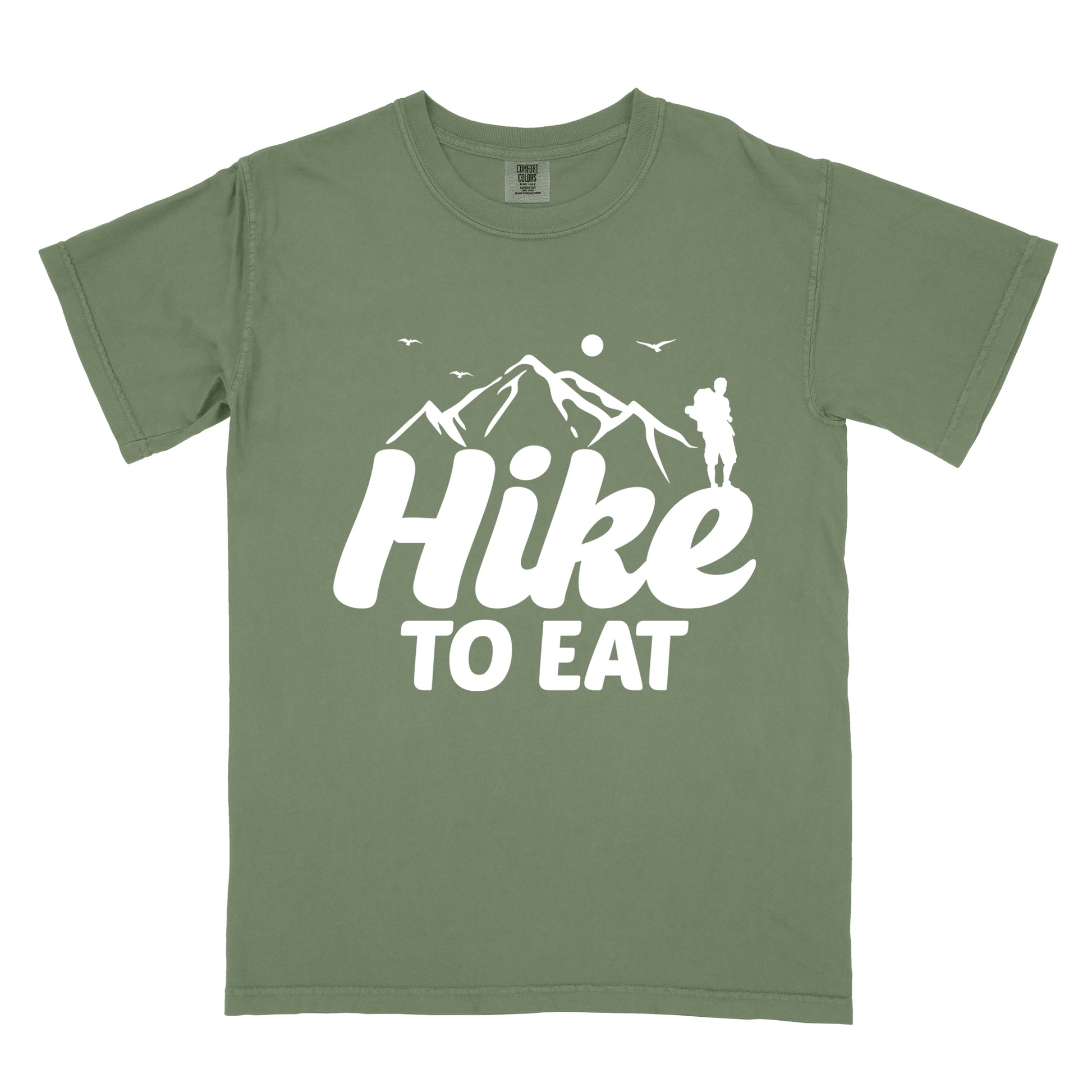 Moss (Dark Green) t-shirt with white text reading "Hike TO EAT". "Hike" appears in large white script letters with white mountain outlines above it, and "TO EAT" in bold white text below it. Features a white silhouette of a hiker to the right of the mountains, white flying birds, and a white circle representing the sun. 