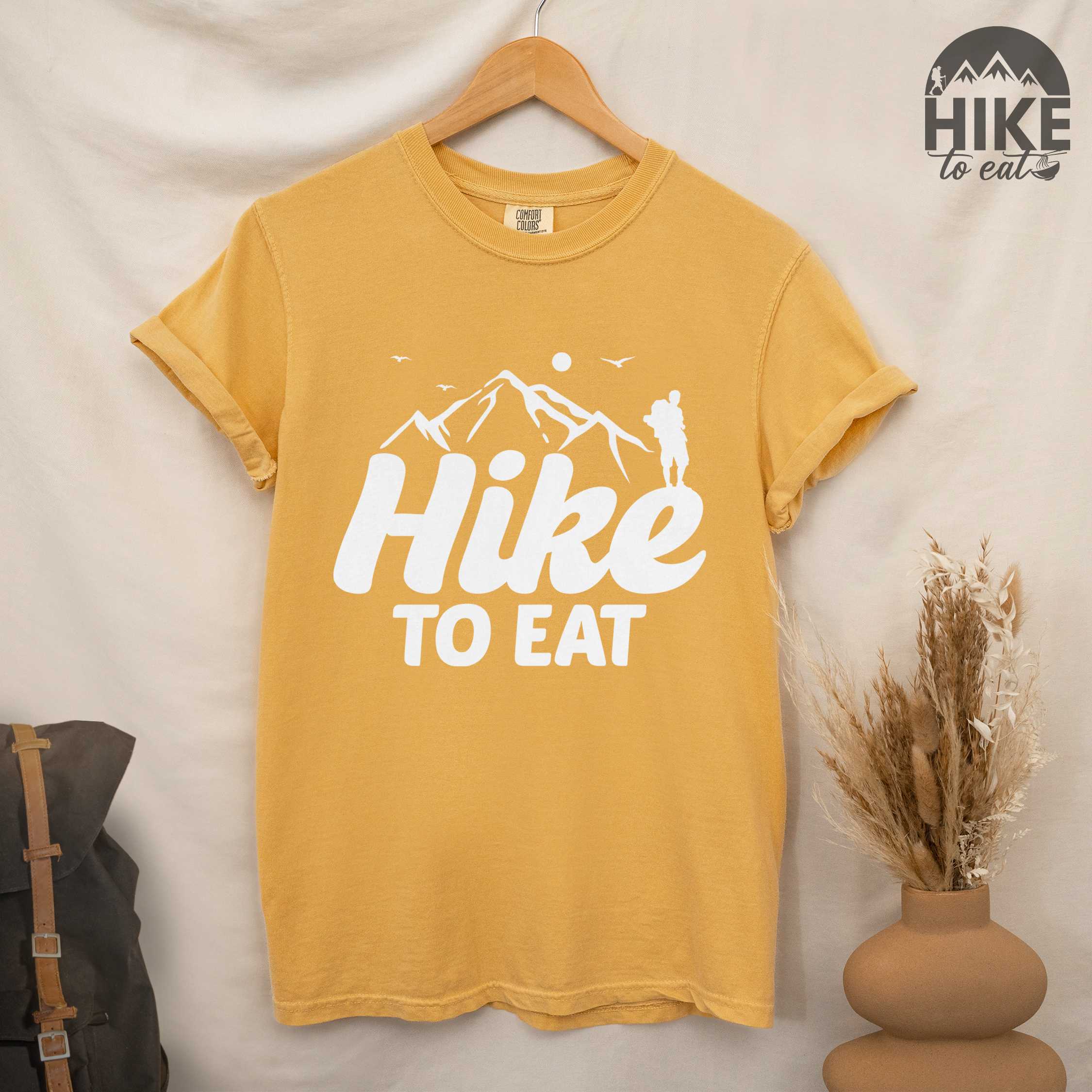 hanging mustard "Hike to Eat" colored statement tee with bold typography, mountains, birds, sunrise, perfect for food motivated individuals.