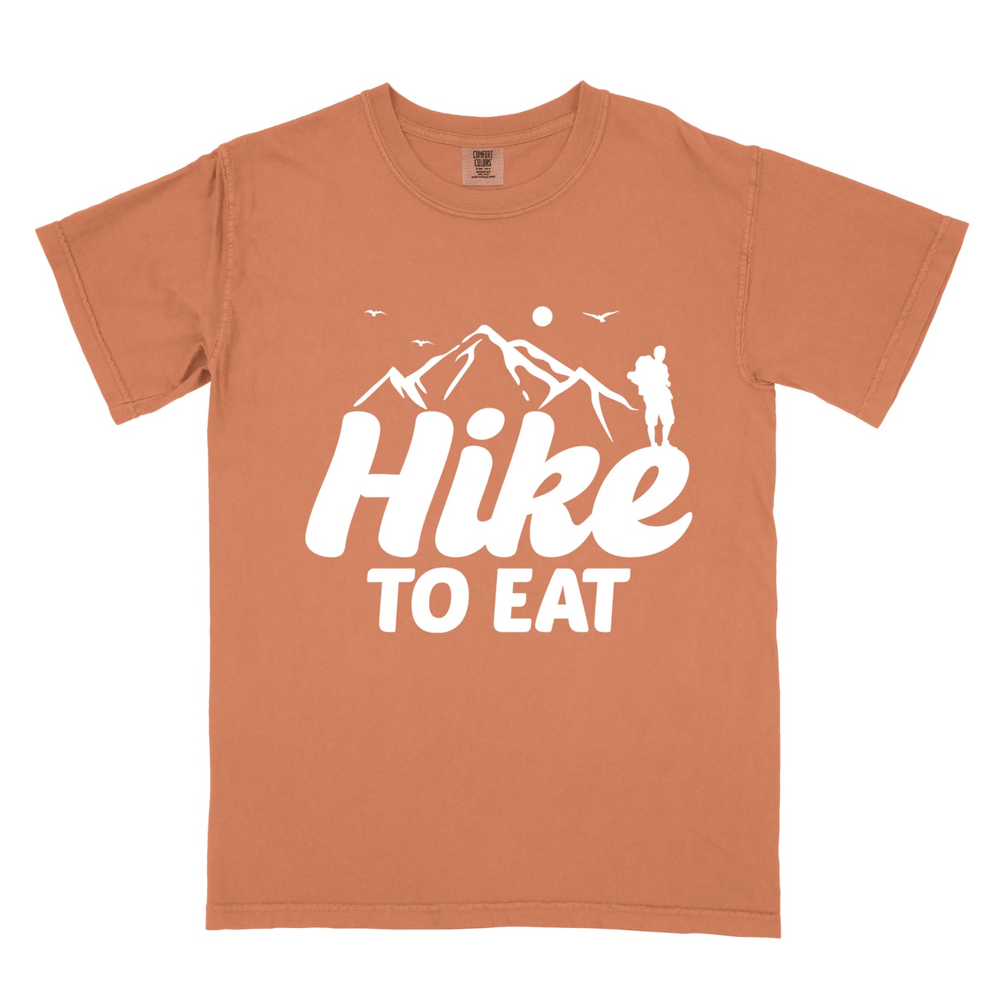 Yam (Brown) t-shirt with white text reading "Hike TO EAT". "Hike" appears in large white script letters with white mountain outlines above it, and "TO EAT" in bold white text below it. Features a white silhouette of a hiker to the right of the mountains, white flying birds, and a white circle representing the sun. 