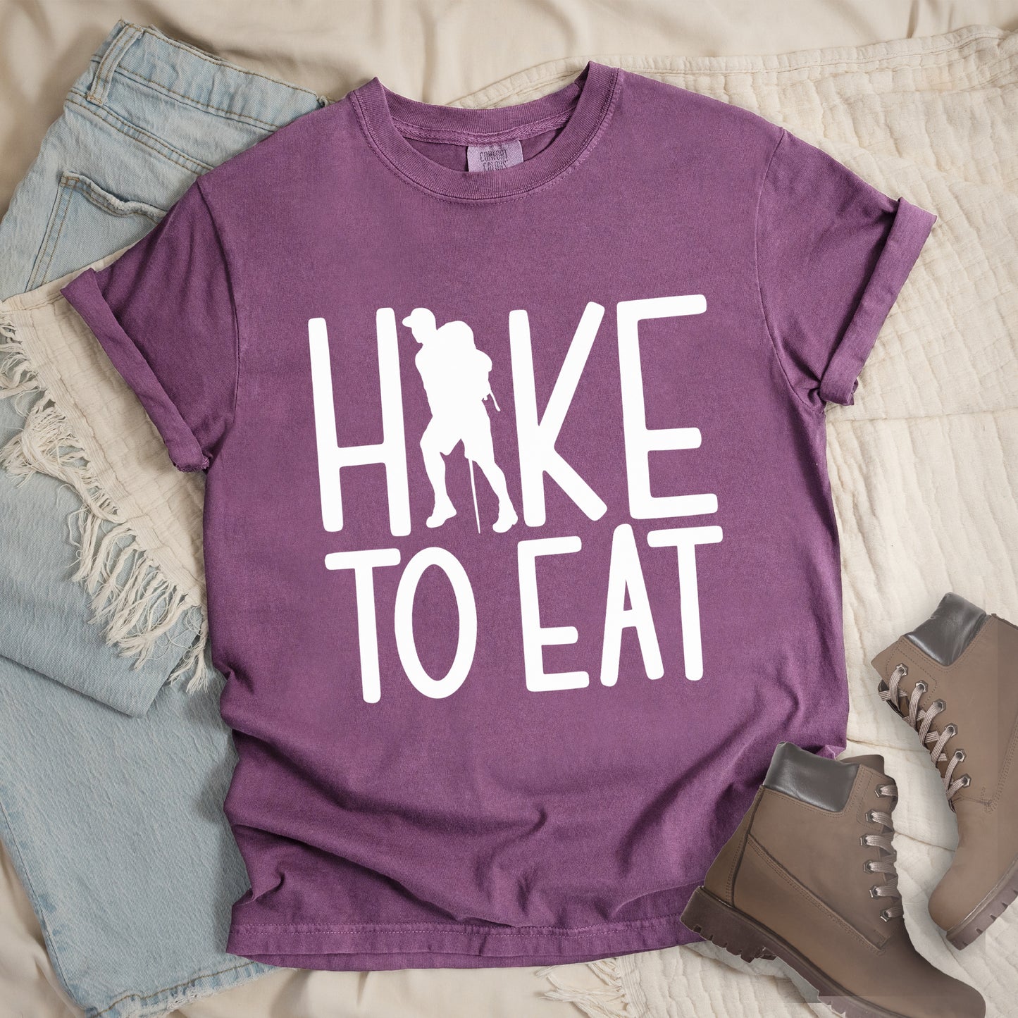 Berry (Dark Violet) t-shirt with white text reading "HIKE TO EAT". "HIKE" appears in large white letters at the top with a white silhouette of a hiker with a backpack and walking stick integrated into the letter "I", and "TO EAT" in white text below it. The shirt features a classic crew neck and casual rolled sleeves, styled alongside light wash denim and brown hiking boots.