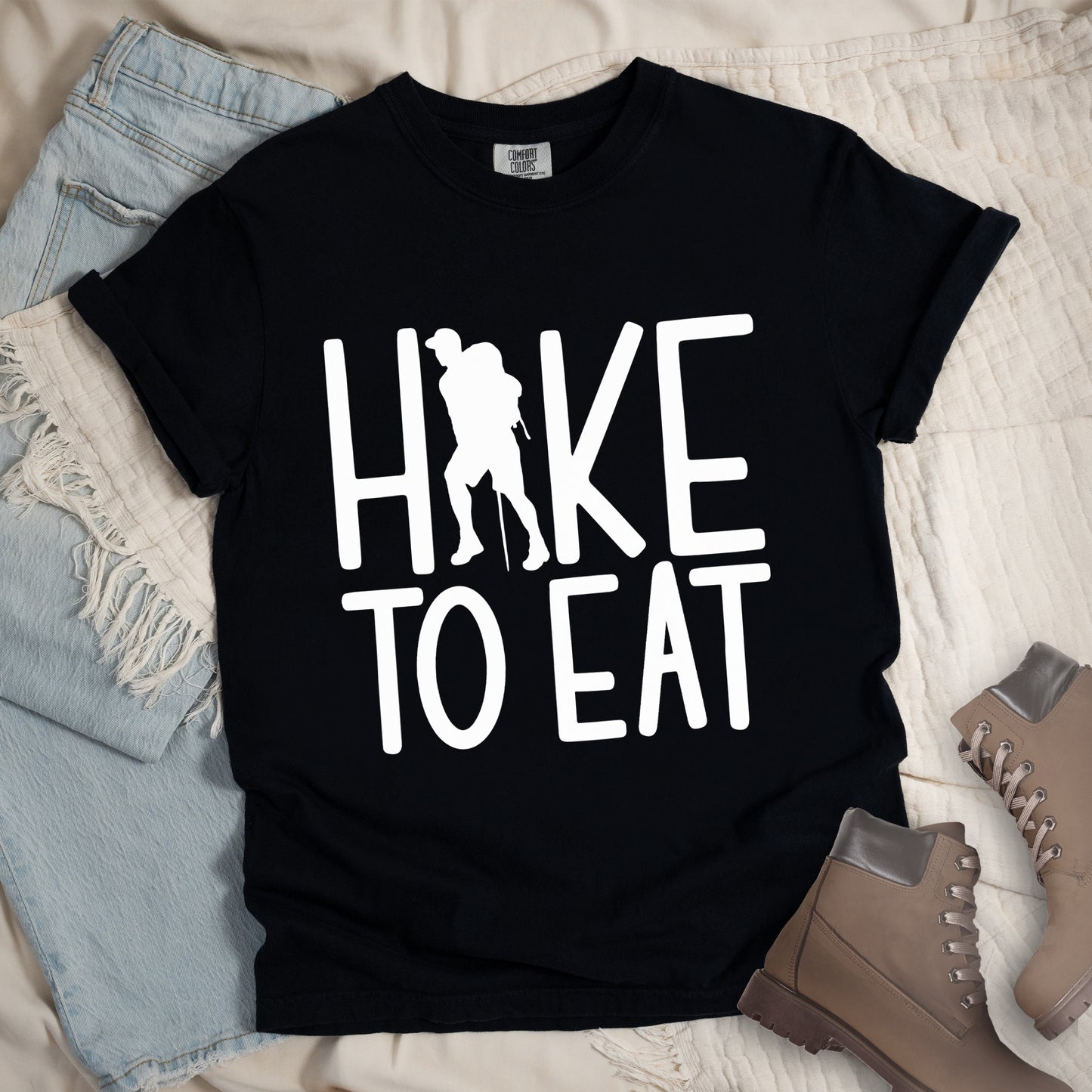 Black t-shirt with white text reading "HIKE TO EAT". "HIKE" appears in large white letters at the top with a white silhouette of a hiker with a backpack and walking stick integrated into the letter "I", and "TO EAT" in white text below it. The shirt features a classic crew neck and casual rolled sleeves, styled alongside light wash denim and brown hiking boots.