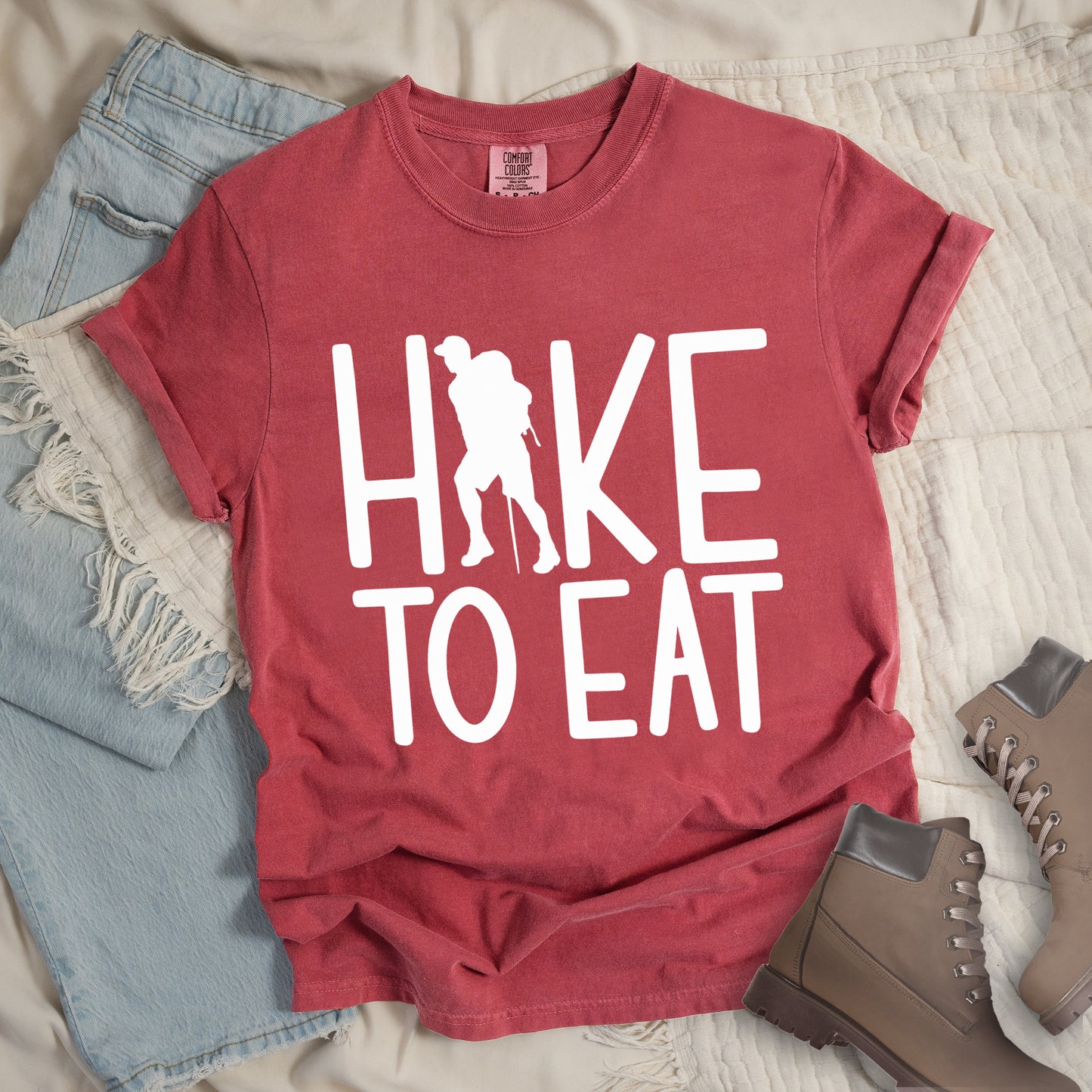 Crimson (Dark Pink) t-shirt with white text reading "HIKE TO EAT". "HIKE" appears in large white letters at the top with a white silhouette of a hiker with a backpack and walking stick integrated into the letter "I", and "TO EAT" in white text below it. The shirt features a classic crew neck and casual rolled sleeves, styled alongside light wash denim and brown hiking boots.