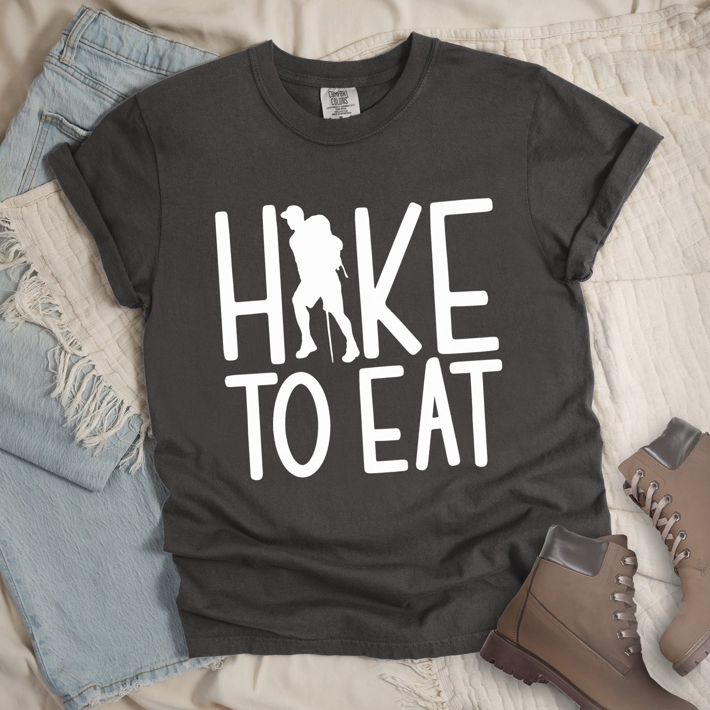 Pepper (Dark Gray) t-shirt with white text reading "HIKE TO EAT". "HIKE" appears in large white letters at the top with a white silhouette of a hiker with a backpack and walking stick integrated into the letter "I", and "TO EAT" in white text below it. The shirt features a classic crew neck and casual rolled sleeves, styled alongside light wash denim and brown hiking boots.