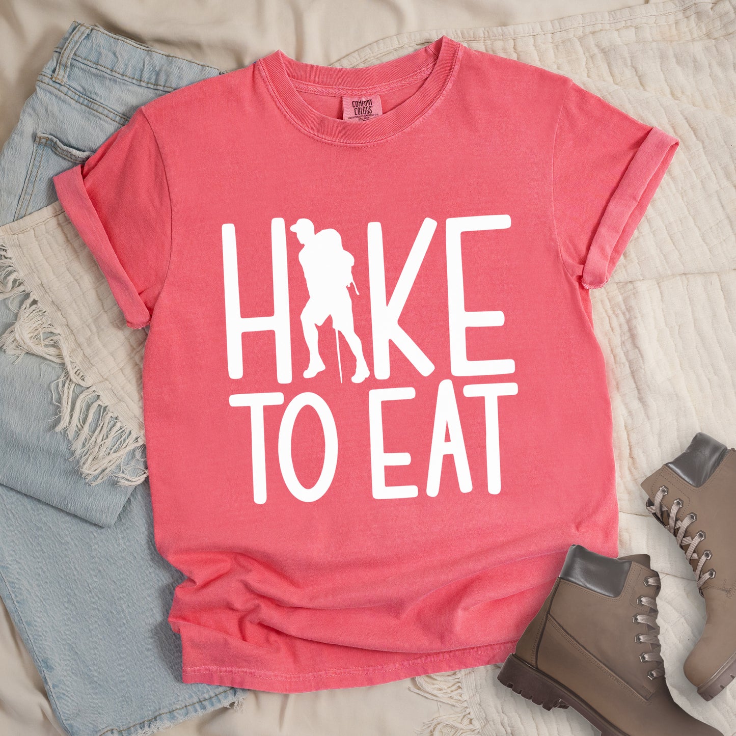 Watermelon (Light Coral) t-shirt with white text reading "HIKE TO EAT". "HIKE" appears in large white letters at the top with a white silhouette of a hiker with a backpack and walking stick integrated into the letter "I", and "TO EAT" in white text below it. The shirt features a classic crew neck and casual rolled sleeves, styled alongside light wash denim and brown hiking boots.