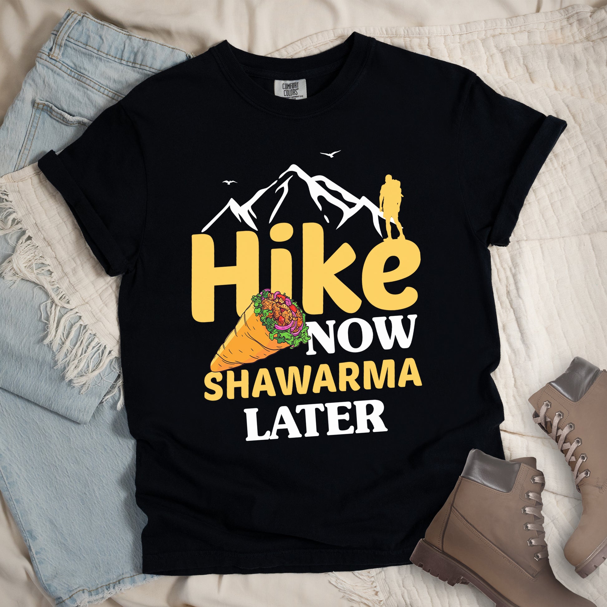 Black t-shirt with text "HIKE NOW SHAWARMA LATER" in yellow and white lettering. A mountain silhouette appears above with a small hiker figure. The word "HIKE" is in large yellow text, "NOW" in smaller white text, "SHAWARMA" in yellow with an illustration of a shawarma wrap beside it, and "LATER" in white. The design emphasizes hiking motivation with food reward. Features a classic crew neck and casual rolled sleeves, styled alongside light wash denim and brown hiking boots.