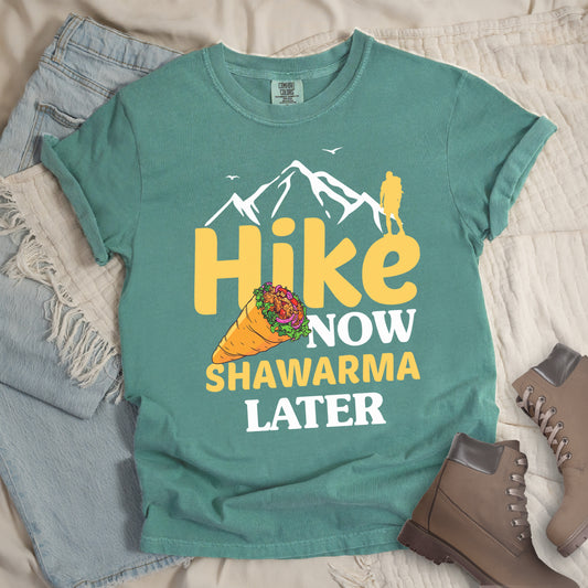 Light Green t-shirt with text "HIKE NOW SHAWARMA LATER" in yellow and white lettering. A mountain silhouette appears above with a small hiker figure. The word "HIKE" is in large yellow text, "NOW" in smaller white text, "SHAWARMA" in yellow with an illustration of a shawarma wrap beside it, and "LATER" in white. The design emphasizes hiking motivation with food reward. Features a classic crew neck and casual rolled sleeves, styled alongside light wash denim and brown hiking boots. 