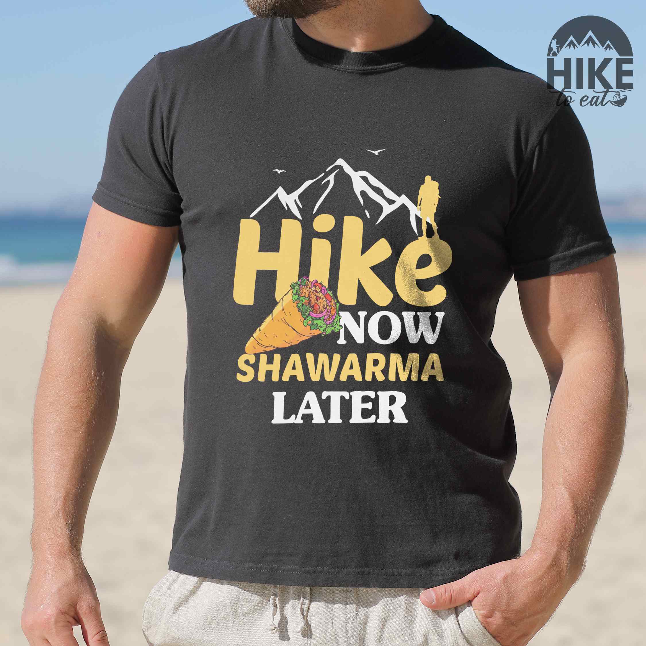 Man wearing black "Hike now Shawarma later" colored vintage tee with bold typography, mountains, birds, and illustrated shawarma. 