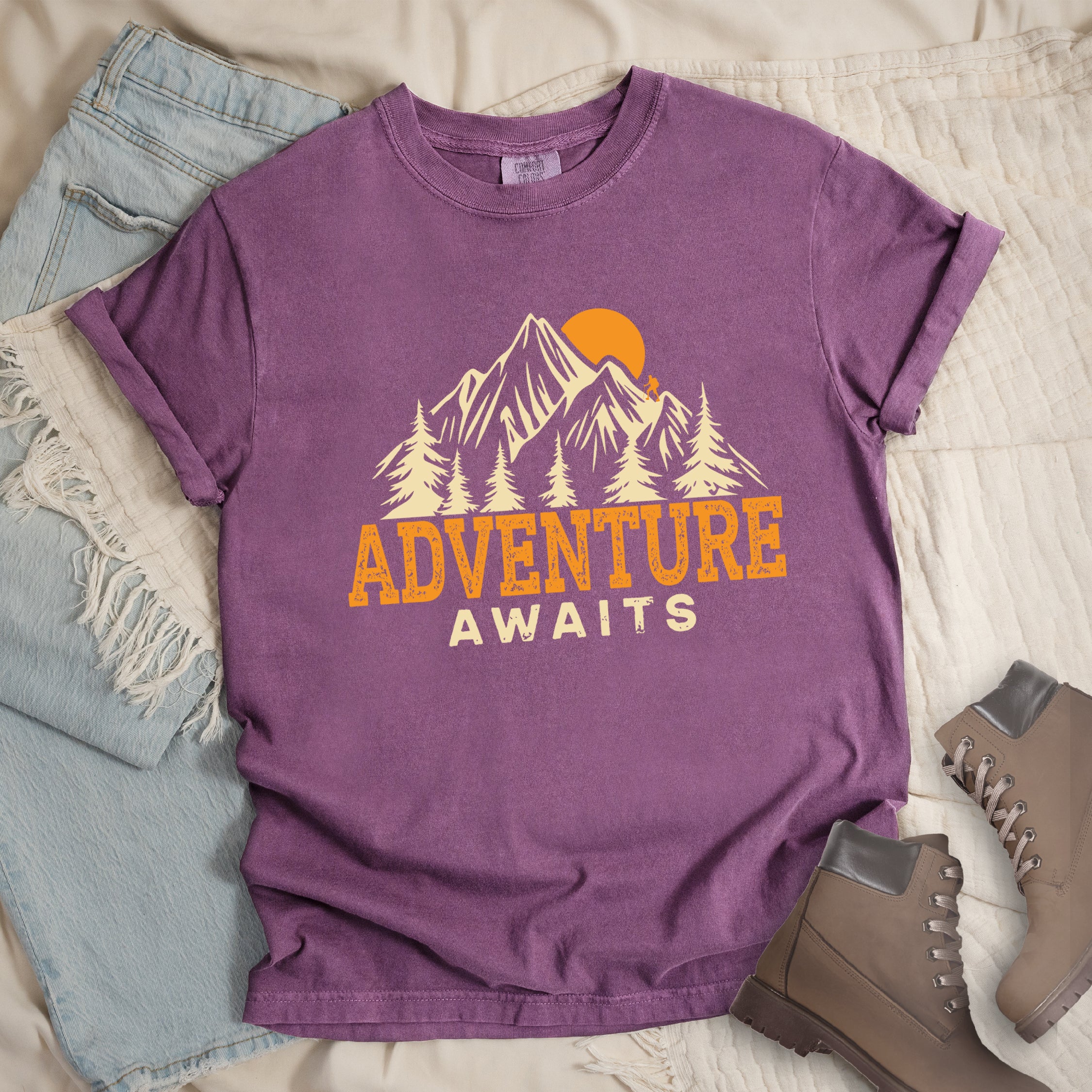 Berry colored (violet) Comfort Colors t-shirt displaying 'Adventure Awaits' graphic with orange distressed lettering, cream-colored mountain peaks, evergreen forest silhouettes, and orange sunset, flat lay styled with faded blue denim jeans and tan lace-up hiking boots on white quilted bedding