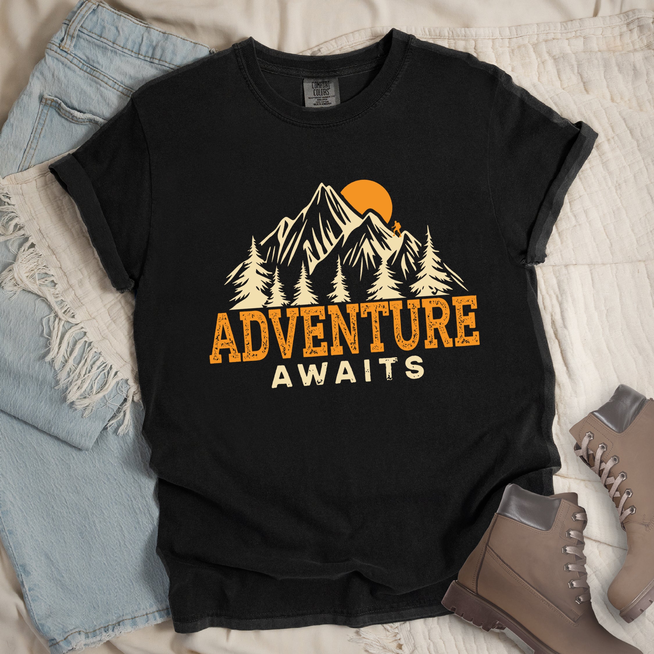 Black Comfort Colors t-shirt displaying 'Adventure Awaits' graphic with orange distressed lettering, cream-colored mountain peaks, evergreen forest silhouettes, and orange sunset, flat lay styled with faded blue denim jeans and tan lace-up hiking boots on white quilted bedding.