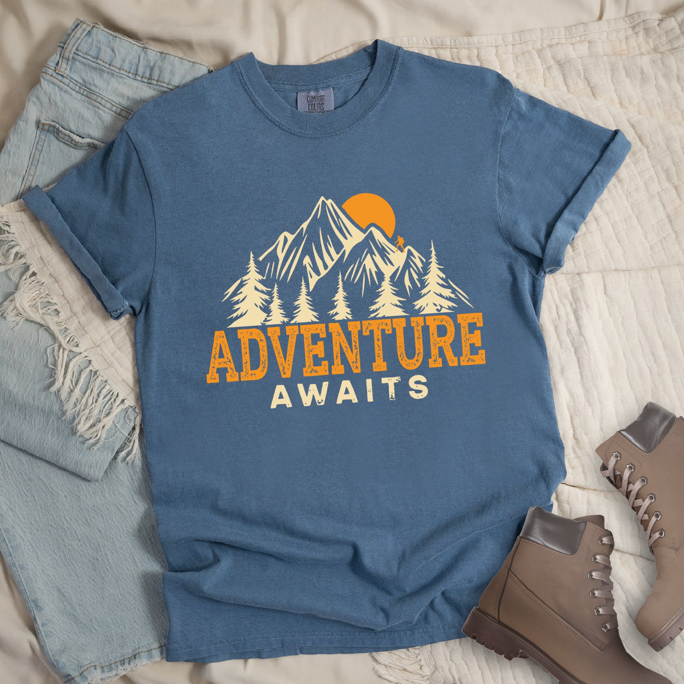 Blue Jean Comfort Colors t-shirt displaying 'Adventure Awaits' graphic with orange distressed lettering, cream-colored mountain peaks, evergreen forest silhouettes, and orange sunset, flat lay styled with faded blue denim jeans and tan lace-up hiking boots on white quilted bedding. 