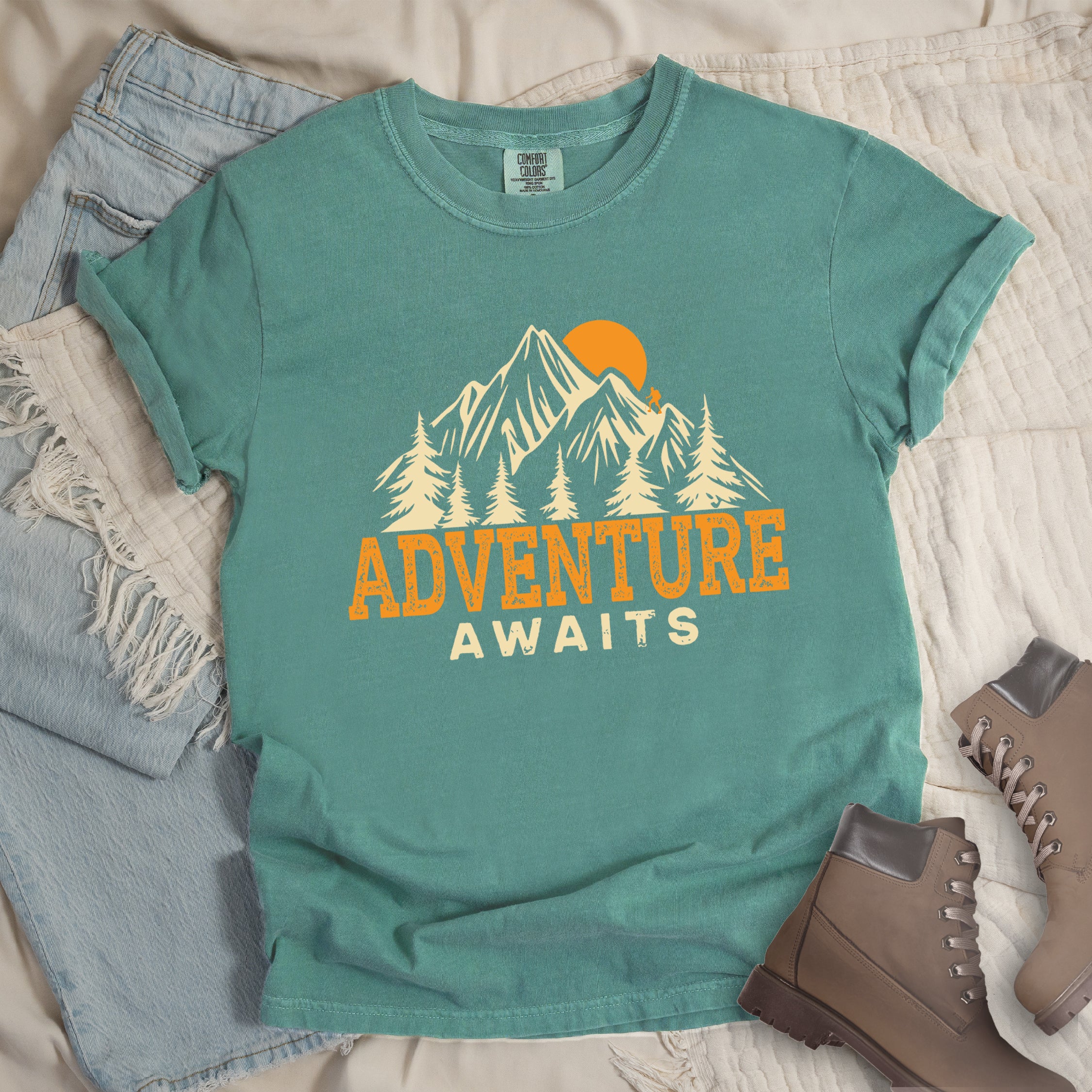 Light Green Comfort Colors t-shirt displaying 'Adventure Awaits' graphic with orange distressed lettering, cream-colored mountain peaks, evergreen forest silhouettes, and orange sunset, flat lay styled with faded blue denim jeans and tan lace-up hiking boots on white quilted bedding