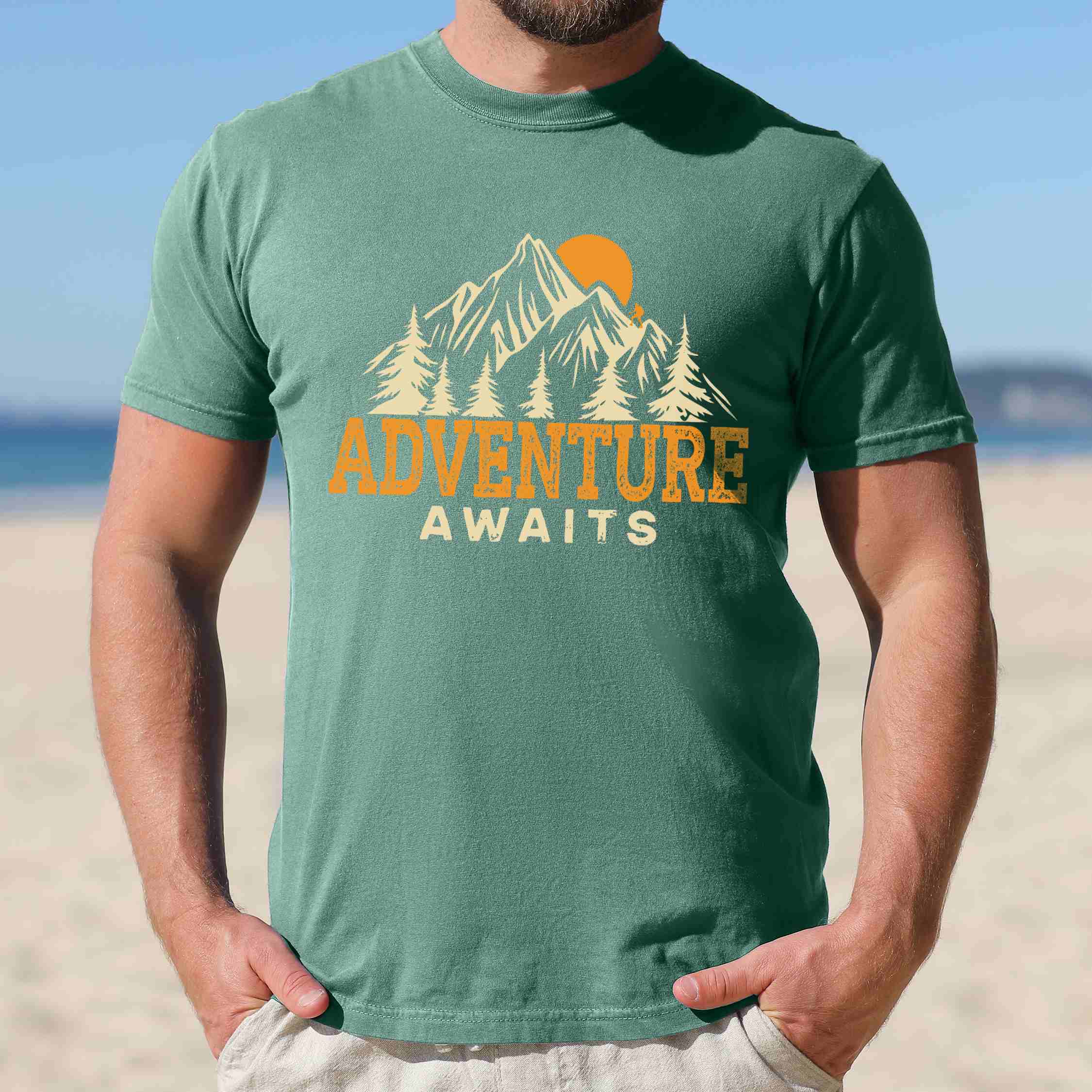 Man wearing light green "Adventure Awaits" colored statement tee with bold typography, mountains, birds, perfect for adventure motivated individuals.