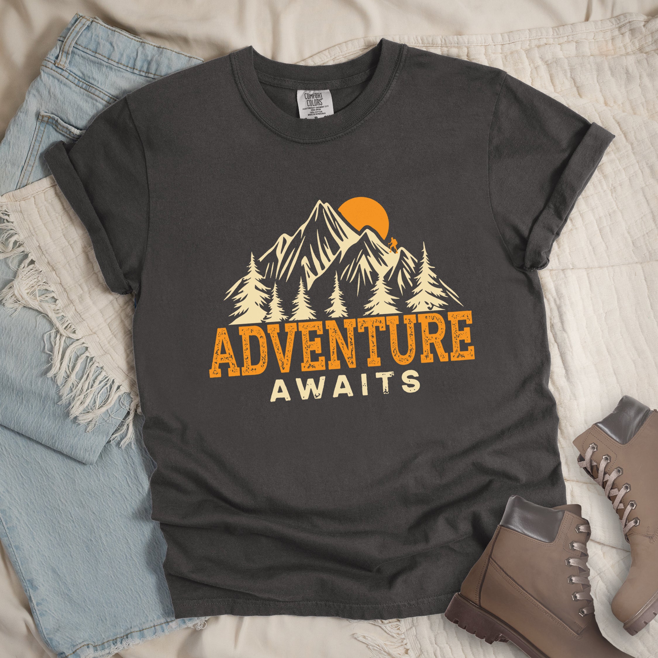 Pepper colored Comfort Colors t-shirt displaying 'Adventure Awaits' graphic with orange distressed lettering, cream-colored mountain peaks, evergreen forest silhouettes, and orange sunset, flat lay styled with faded blue denim jeans and tan lace-up hiking boots on white quilted bedding