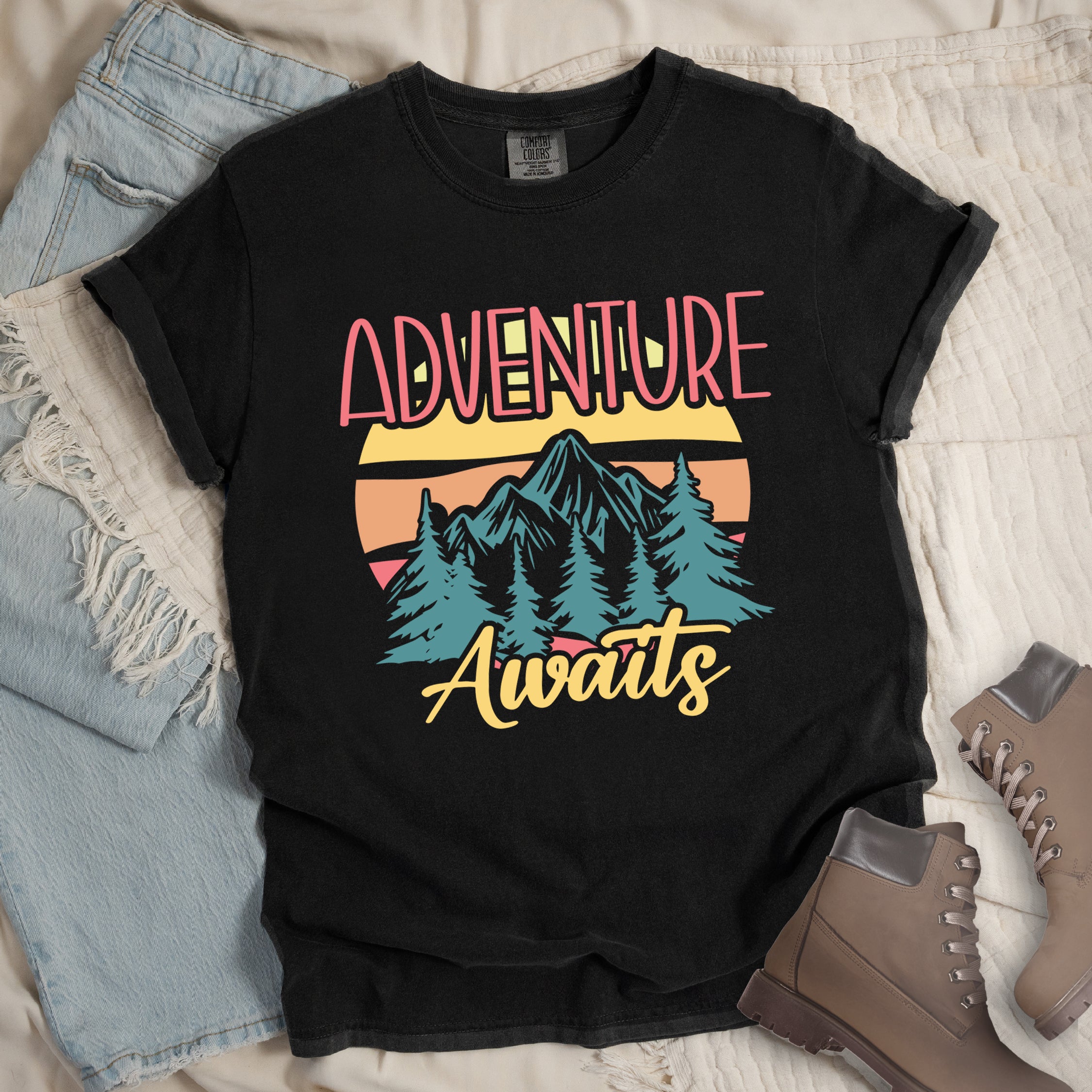 Black t-shirt with retro-style 'Adventure Awaits' design featuring pink gradient text above mountain landscape with teal pine trees, colorful sunset stripes in yellow, orange and pink, and yellow script 'Awaits' text below. 