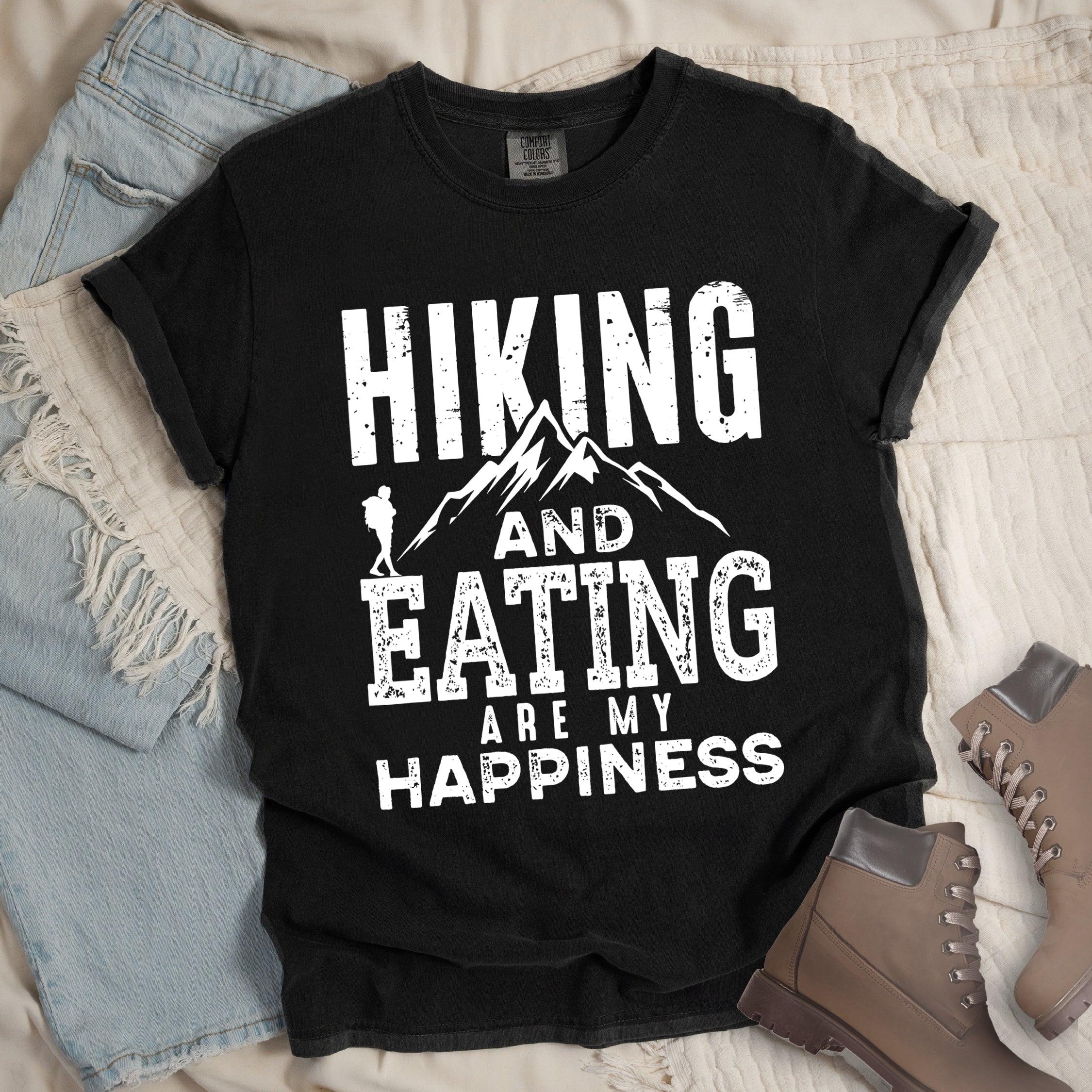 Black colored t-shirt with white distressed text reading 'Hiking and Eating Are My Happiness' featuring mountain silhouette and hiker figure, styled with light wash denim jeans and brown hiking boots on cream bedding.