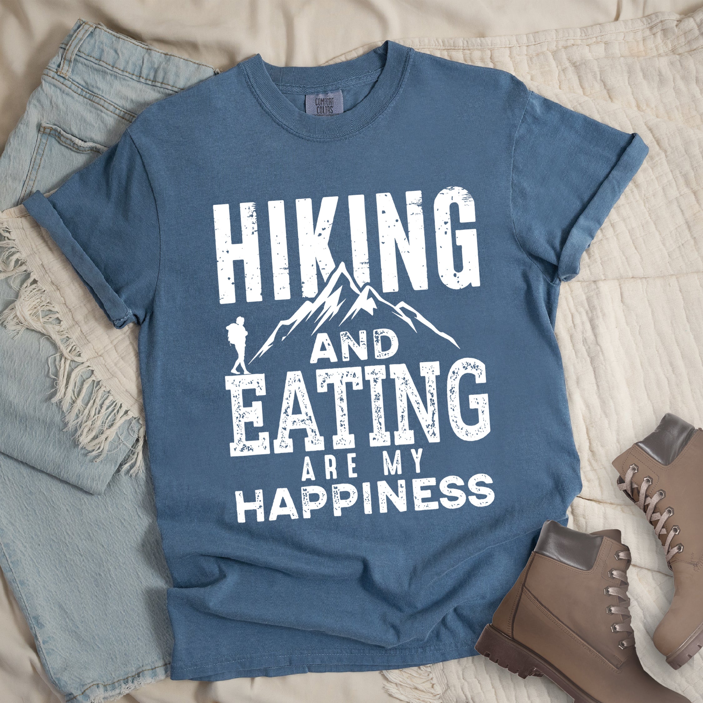 Blue jean colored t-shirt with white distressed text reading 'Hiking and Eating Are My Happiness' featuring mountain silhouette and hiker figure, styled with light wash denim jeans and brown hiking boots on cream bedding.