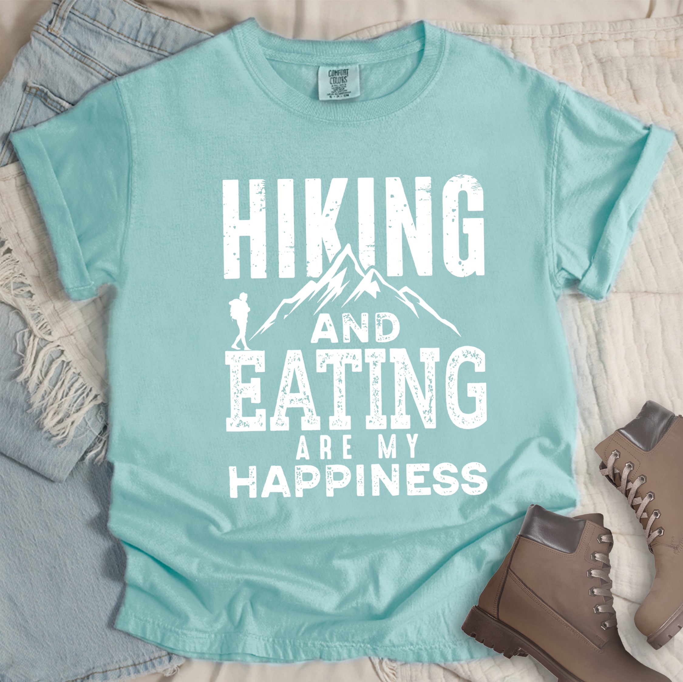 Chalky mint colored t-shirt with white distressed text reading 'Hiking and Eating Are My Happiness' featuring mountain silhouette and hiker figure, styled with light wash denim jeans and brown hiking boots on cream bedding.