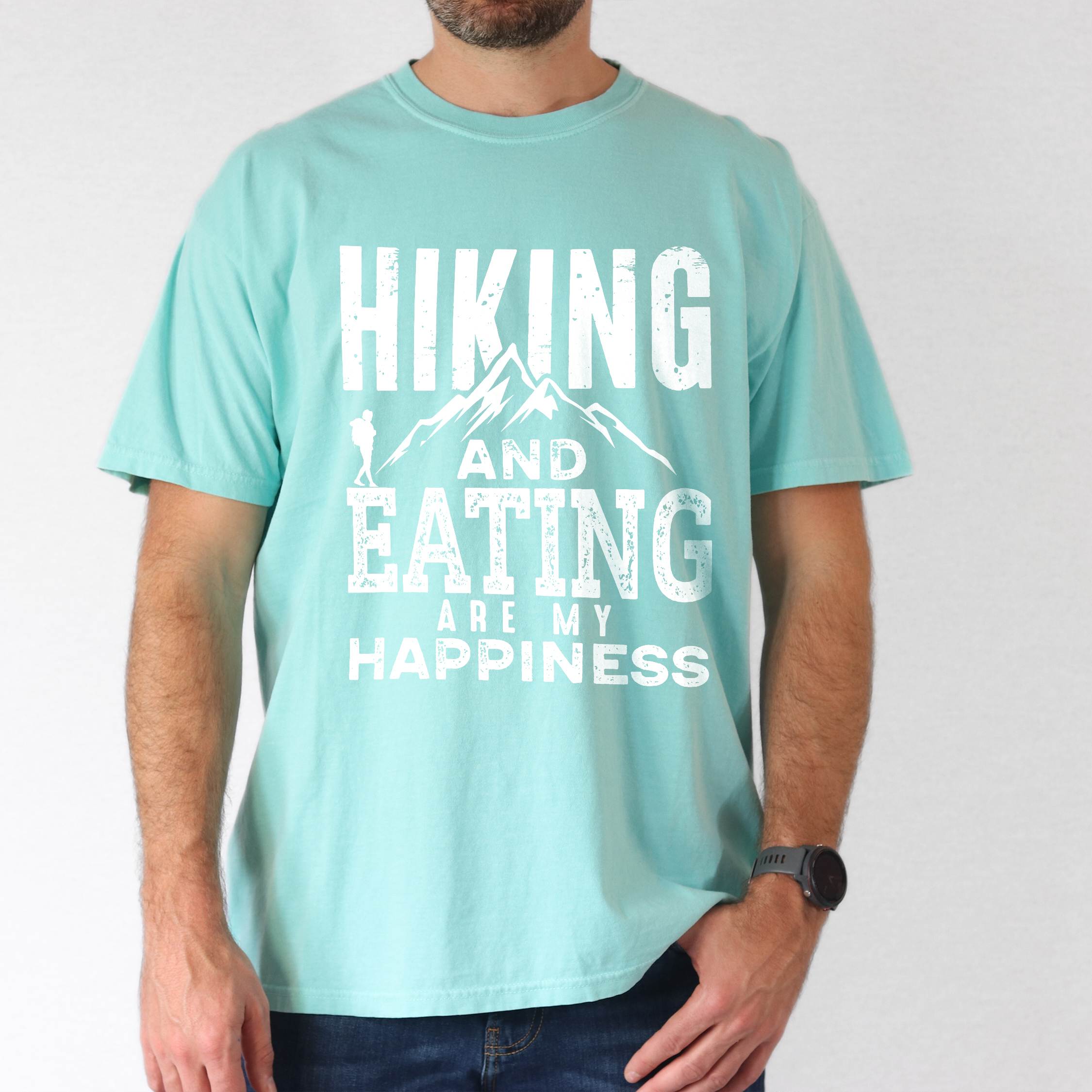 Man wearing chalky mint "Hiking and Eating are my happiness" colored statement tee with bold typography, mountains, birds, perfect for adventure motivated individuals.