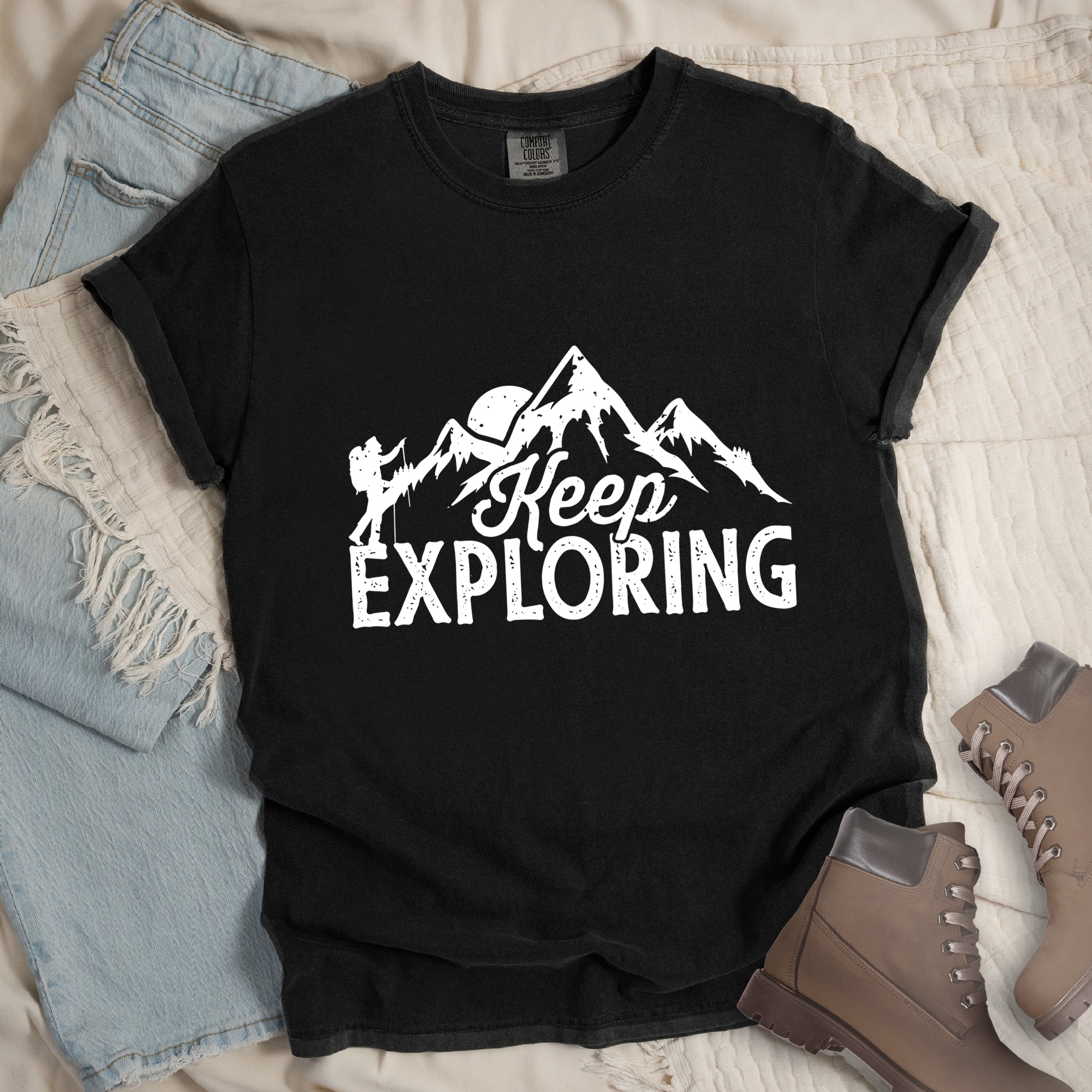 Black colored t-shirt with white distressed 'Keep Exploring' text and mountain range silhouette featuring hiker figure and moon, styled with light wash denim jeans and brown hiking boots on cream bedding. 