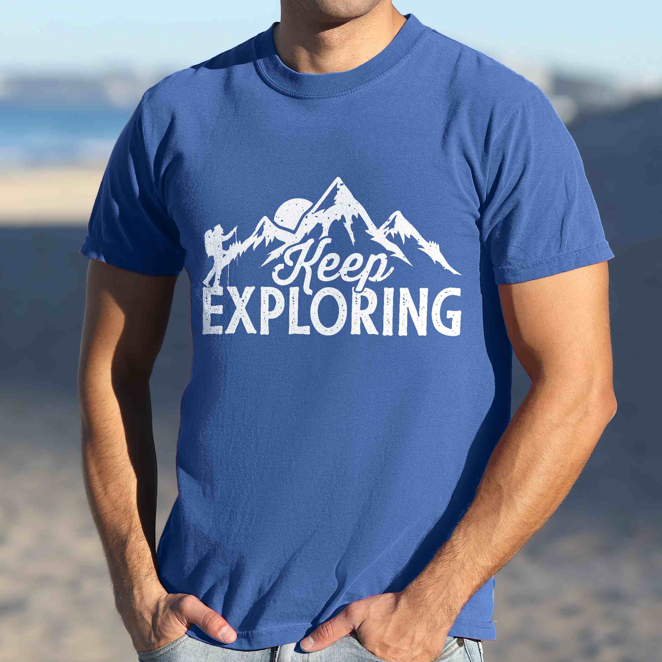 Man wearing flo blue "Keep Exploring" colored statement tee with bold typography, mountains, birds, perfect for adventure motivated individuals.