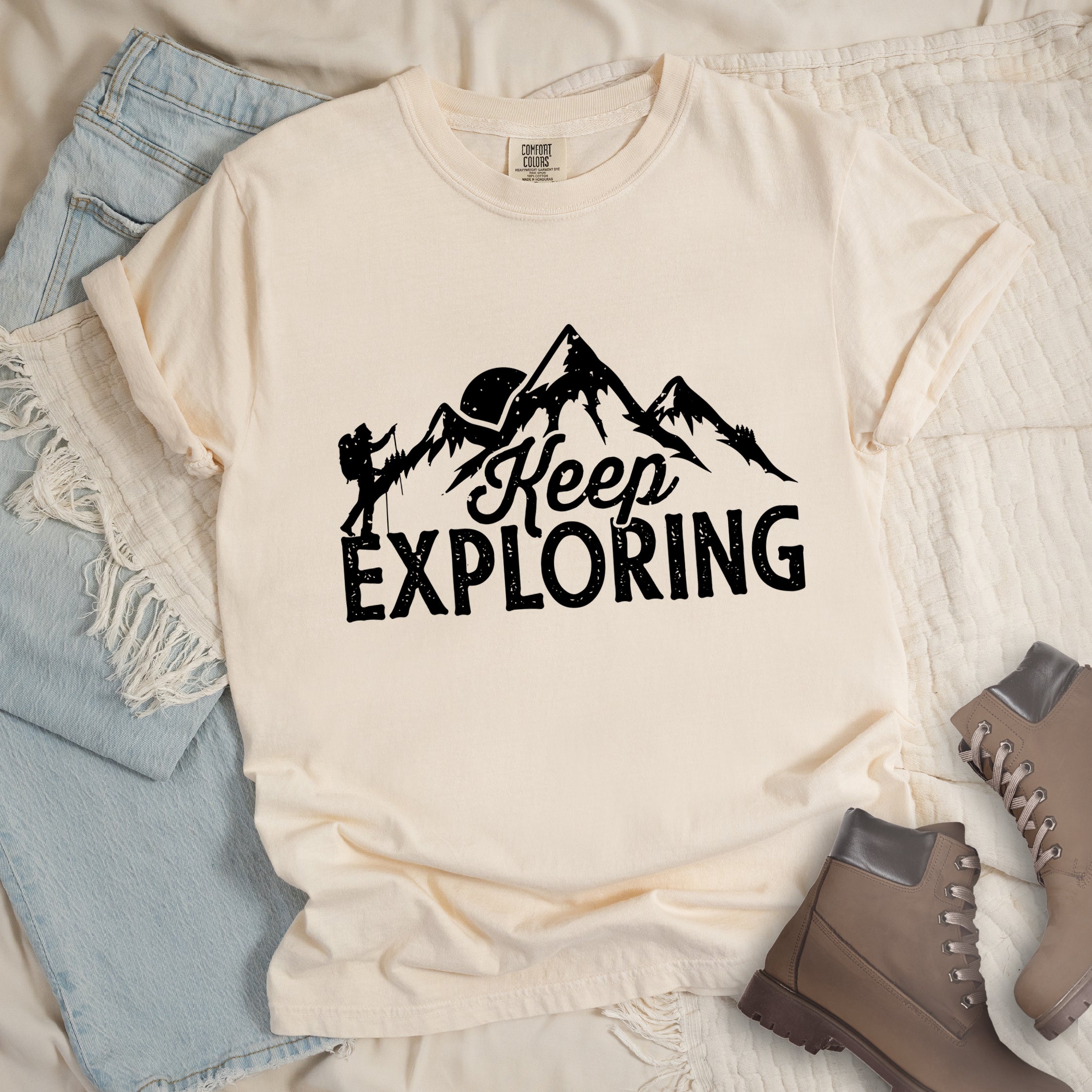 Ivory colored t-shirt with black distressed 'Keep Exploring' text and mountain range silhouette featuring hiker figure and moon, styled with light wash denim jeans and brown hiking boots on cream bedding. 