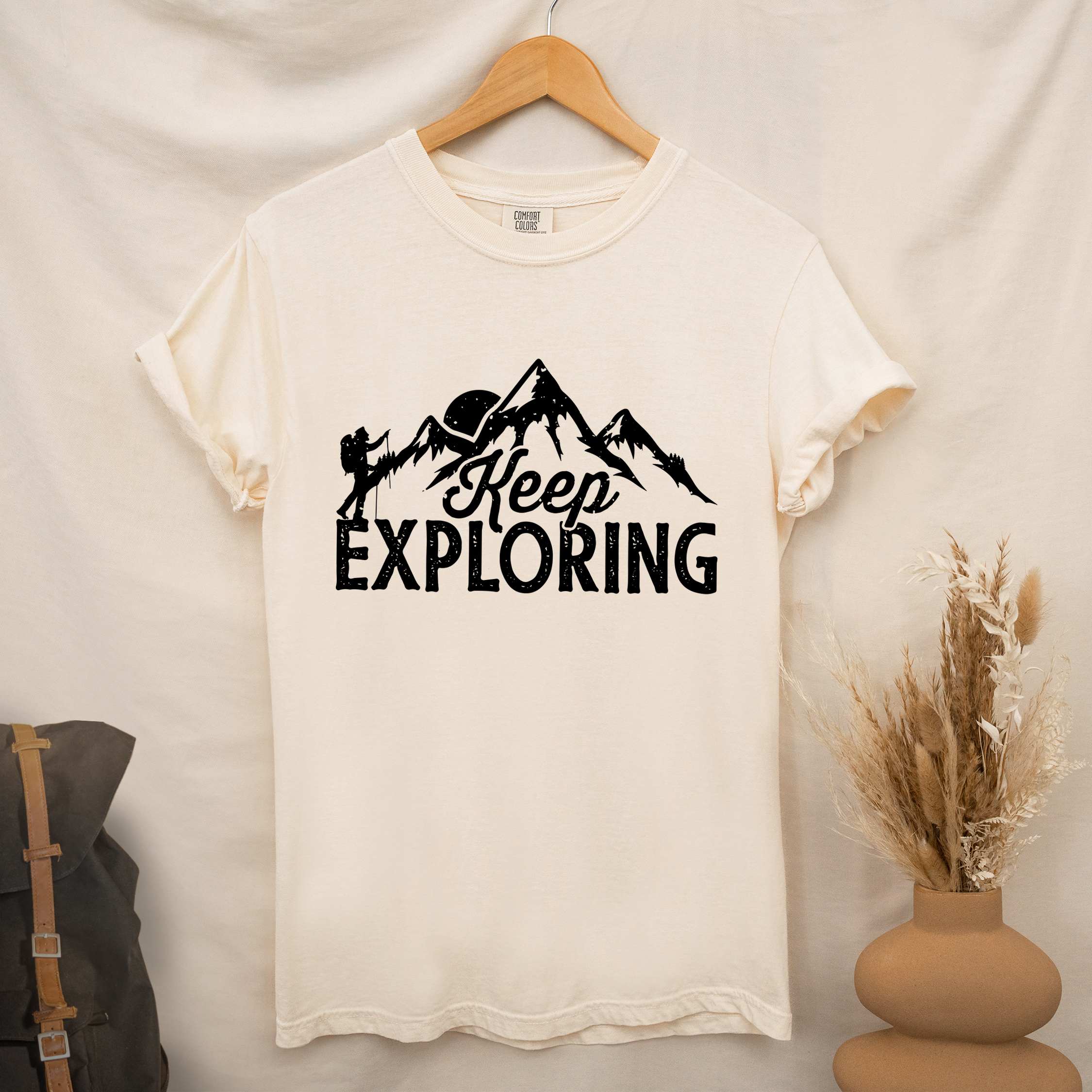 Hanging ivory "Keep Exploring" colored statement tee with bold typography, mountains, birds, perfect for adventure motivated individuals.