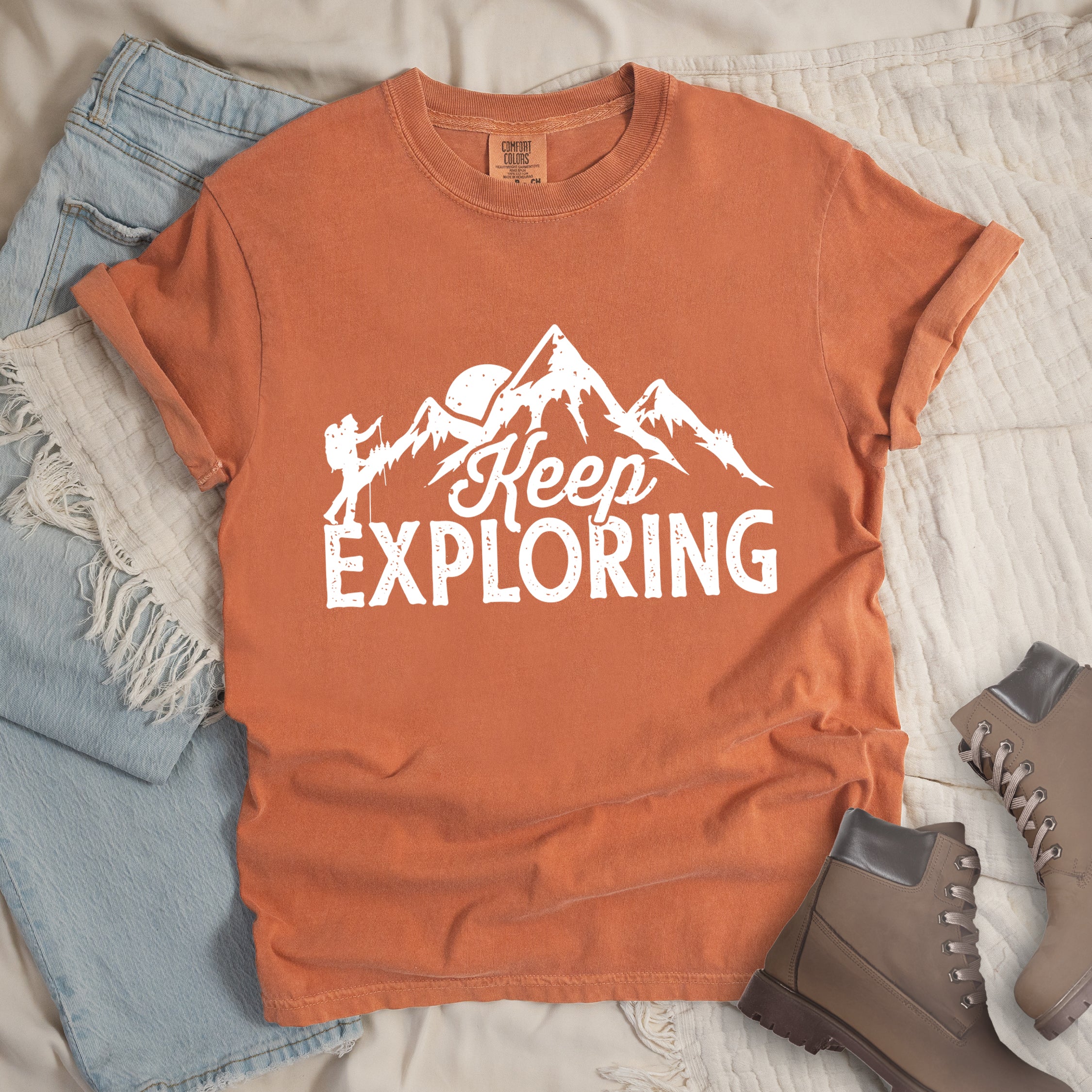 Yam colored t-shirt with white distressed 'Keep Exploring' text and mountain range silhouette featuring hiker figure and moon, styled with light wash denim jeans and brown hiking boots on cream bedding. 