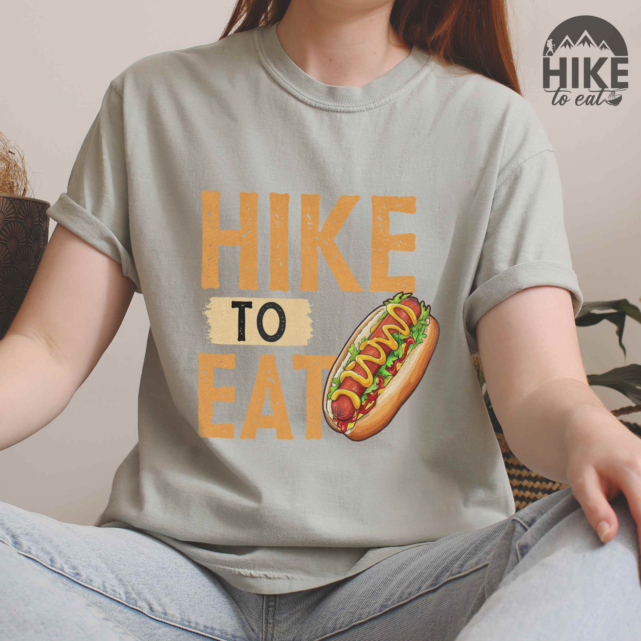 woman wearing bay "Hike to Eat" colored vintage tee with bold typography, and illustrated hotdog with mustard and ketchup sauce.